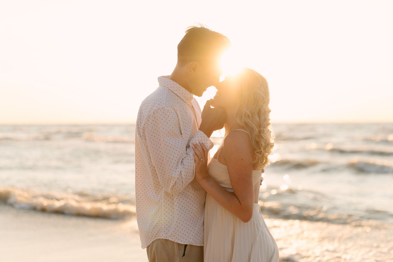 Romantic couple embracing on beach at sunset with golden sun glowing between them by the ocean.