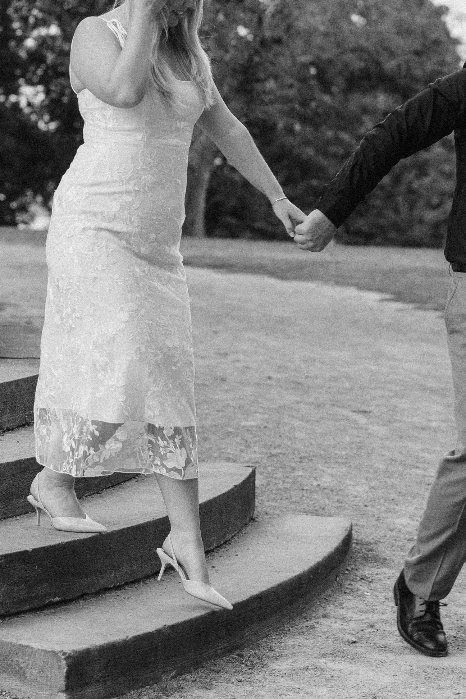 Black and white photo of woman in lace dress descending stone steps while holding man's hand.