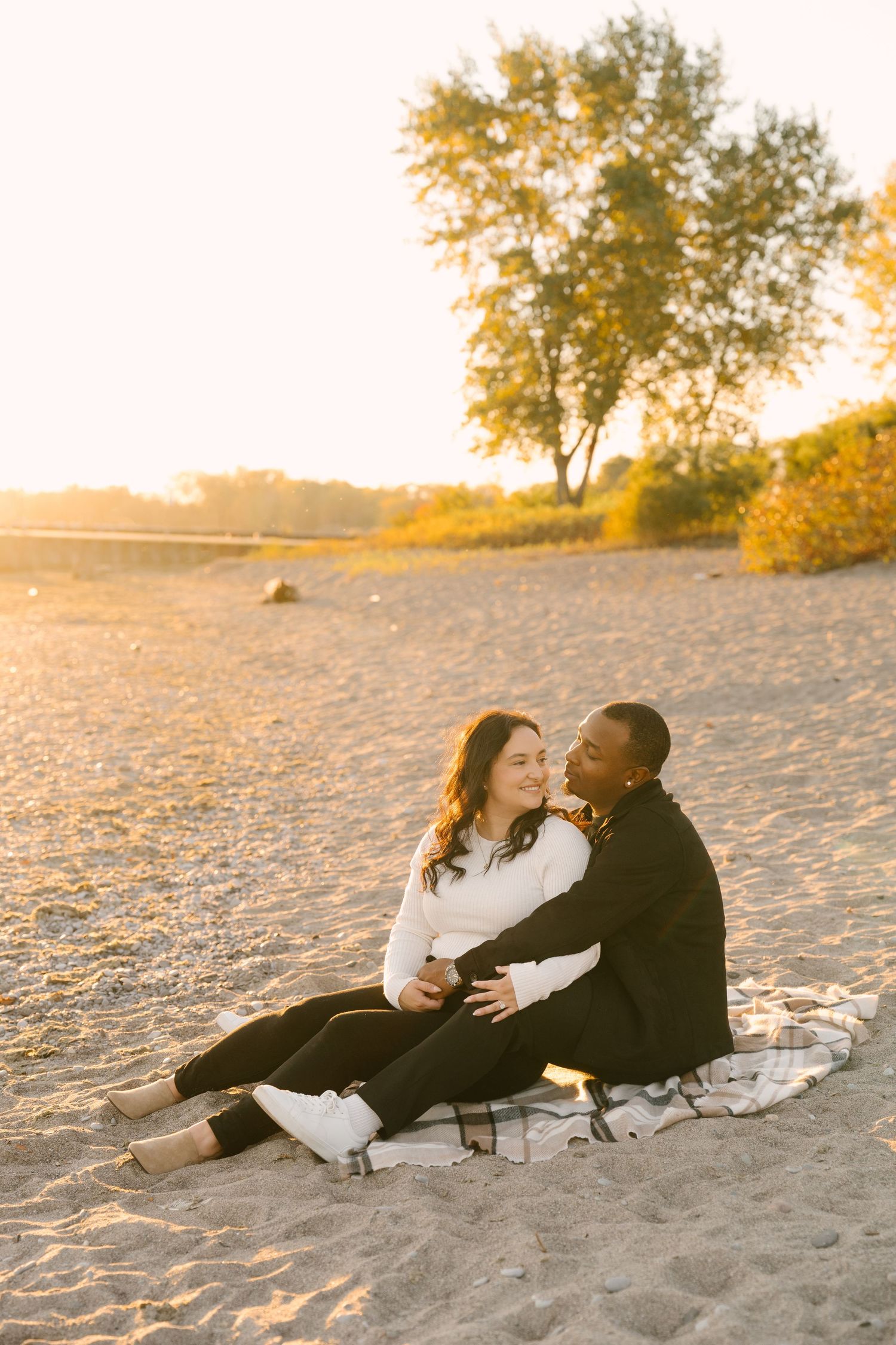 Couple sitting together on a sandy beach blanket at sunset, embracing and smiling.