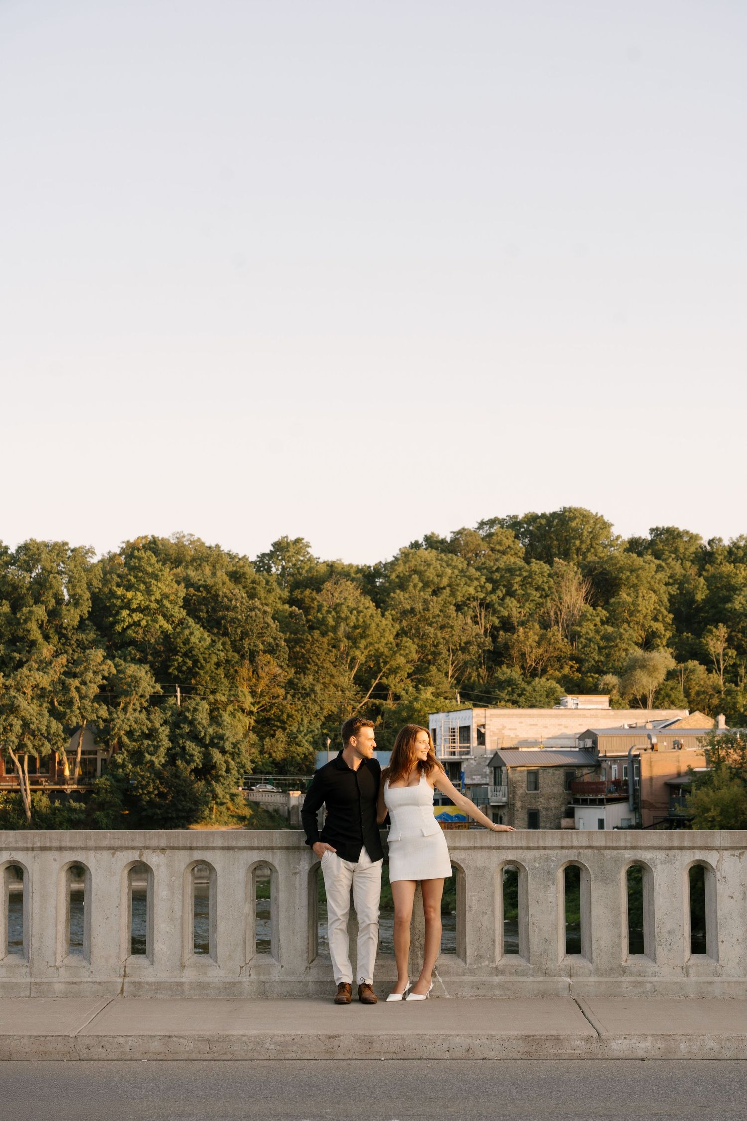 Couple standing together on a stone bridge overlooking lush green trees at golden hour.