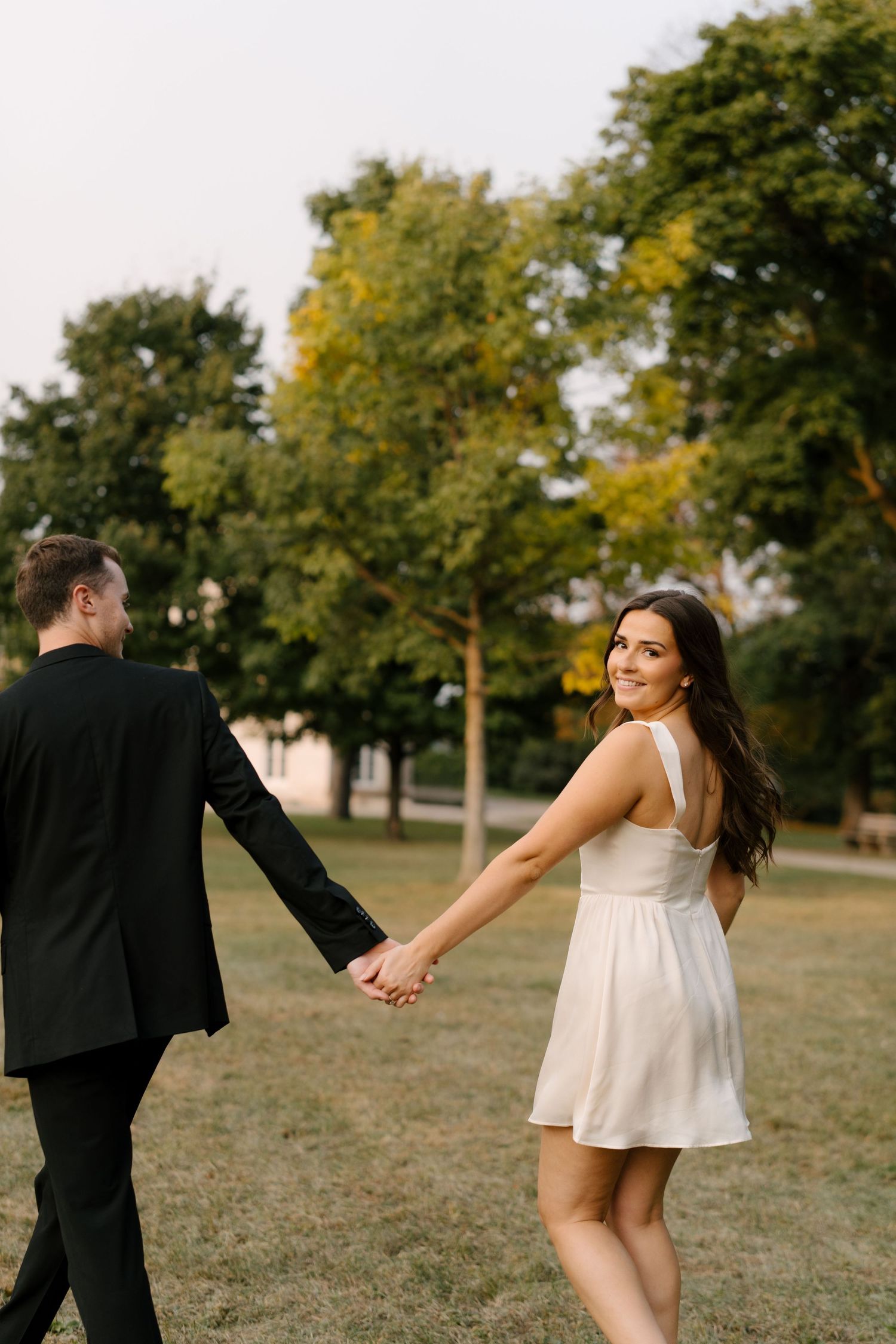 Woman in white dress looks back smiling while holding hands with man in black suit.