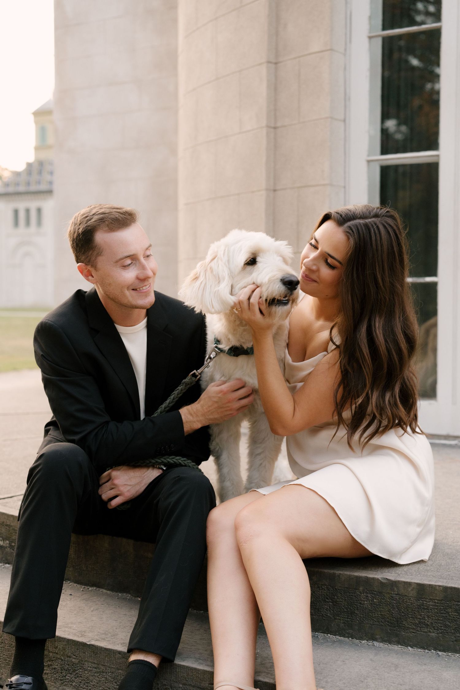 Couple sitting on stone steps with a fluffy white dog at a classic building.