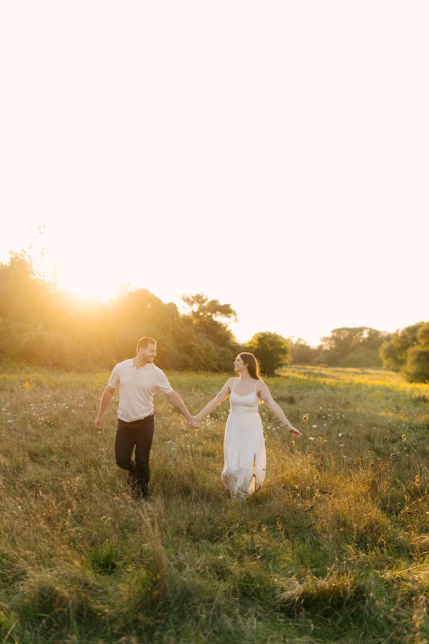 Couple holding hands walking through golden sunlit meadow at sunset, woman in white dress, man in white shirt.