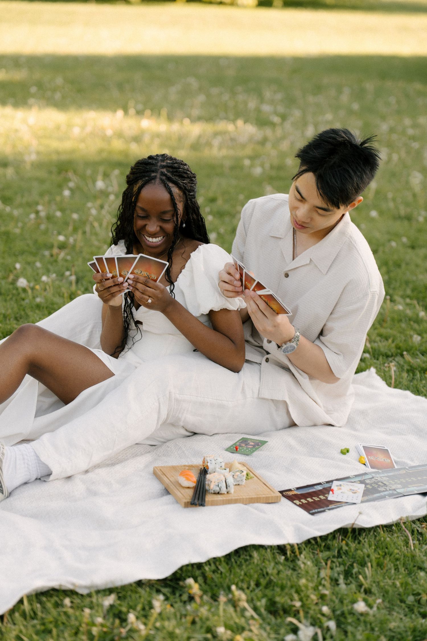 Interracial couple playing cards on a white blanket in a sunny park, smiling and enjoying a romantic outdoor picnic.