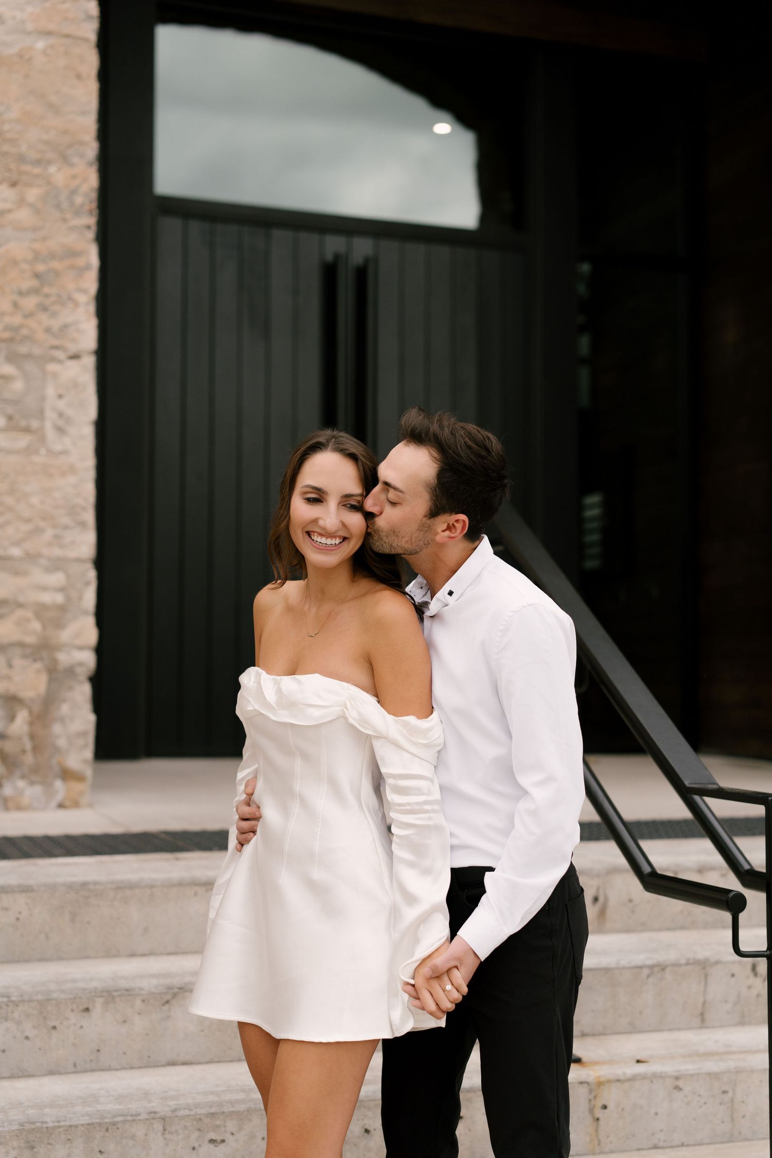 Smiling woman in white mini dress receiving a kiss on the cheek from her partner on stone steps.