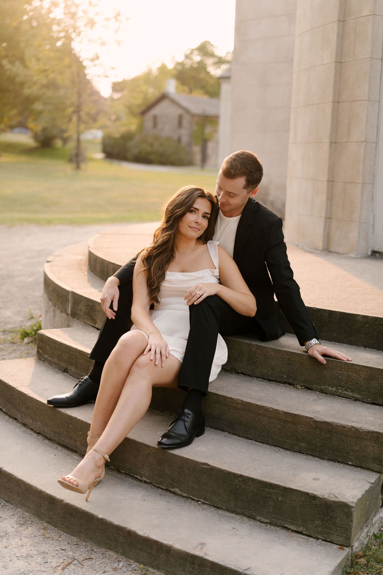 Couple sitting intimately on curved stone steps at golden hour, woman in white mini dress, man in black suit.