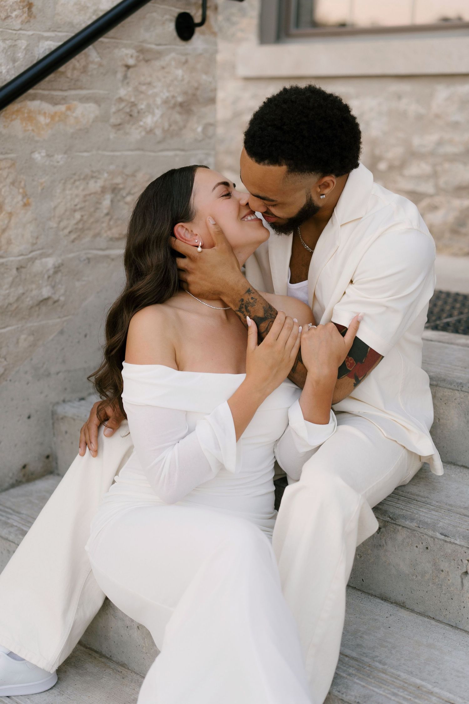 Couple in white outfits sharing romantic kiss while sitting on stone steps during engagement session.