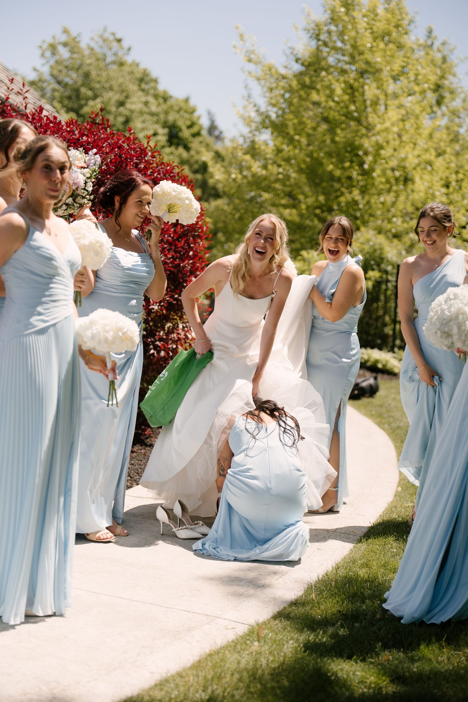 Bride laughing with bridesmaids in light blue gowns holding white bouquets in a garden setting at Roseville Estates wedding venue.