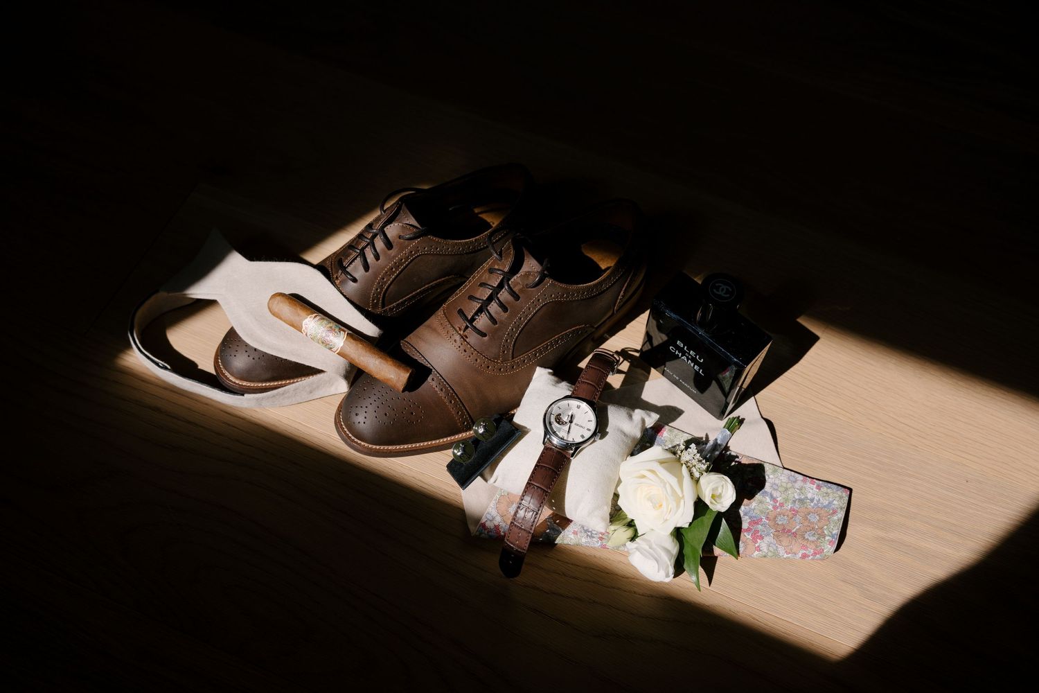 Brown leather dress shoes, watch, boutonniere, and wedding accessories arranged elegantly on a wooden surface.