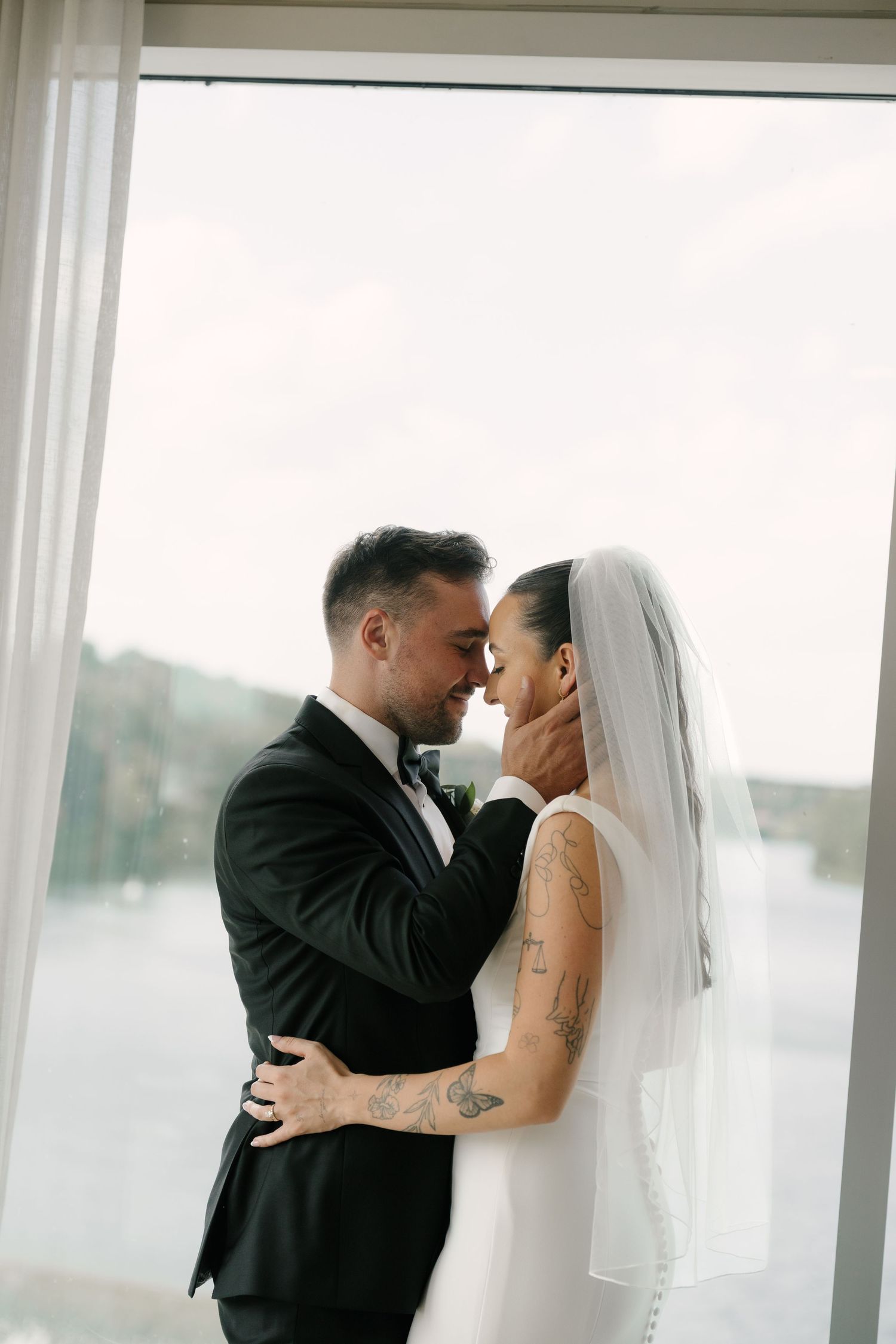 Bride and groom share an intimate moment, touching foreheads by a large window, both with tattoos, she in a veil at Cambridge Mill.
