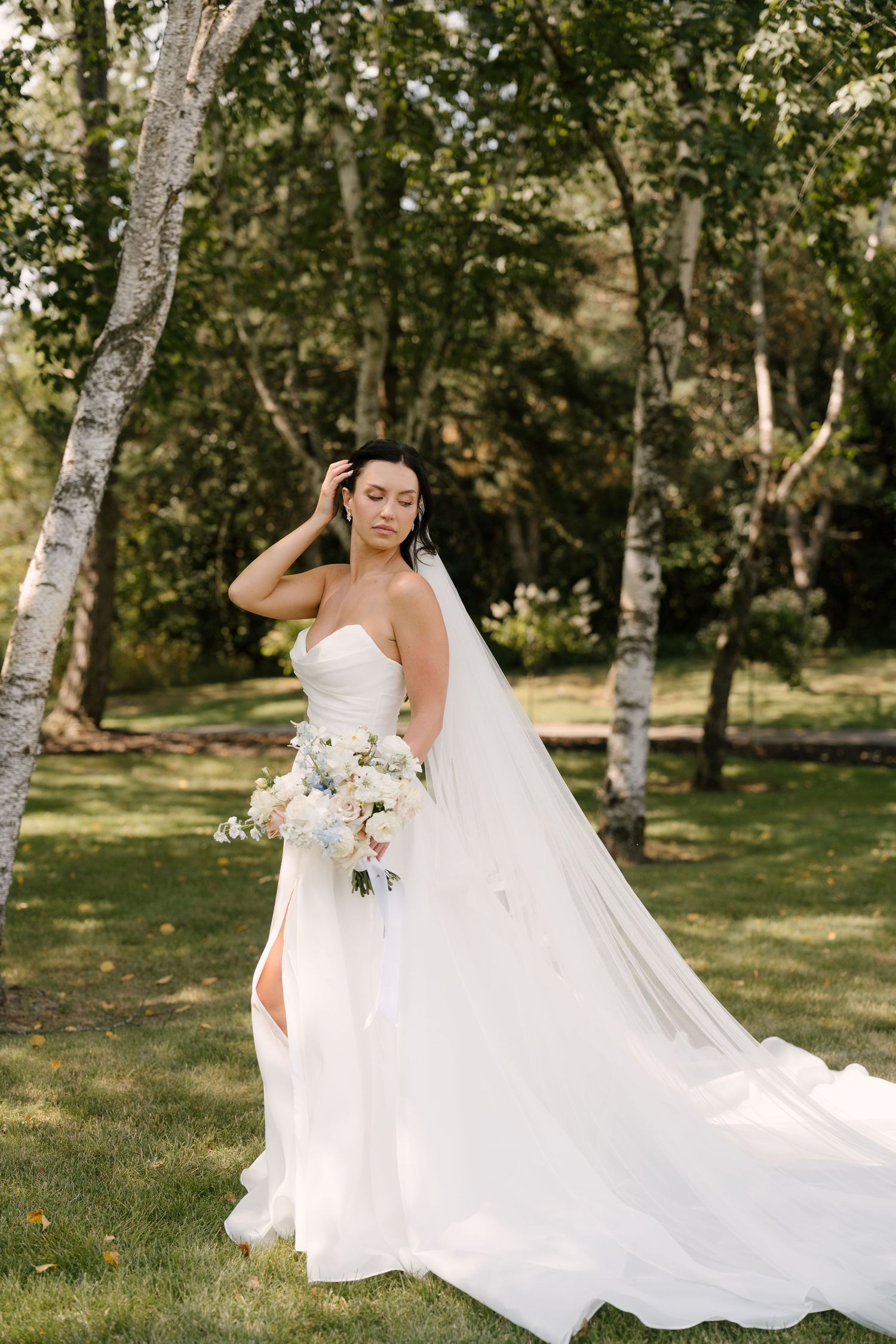 Elegant bride in strapless white gown holding bouquet among birch trees outdoors at Roseville Estates wedding venue.