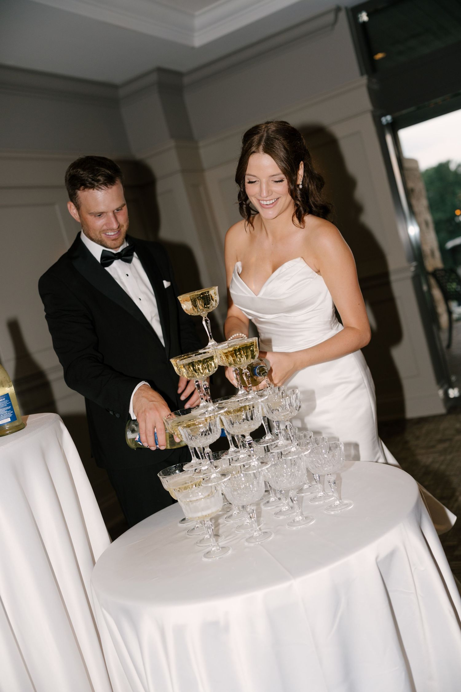 Bride and groom smile while pouring champagne tower at elegant wedding reception with crystal coupe glasses.
