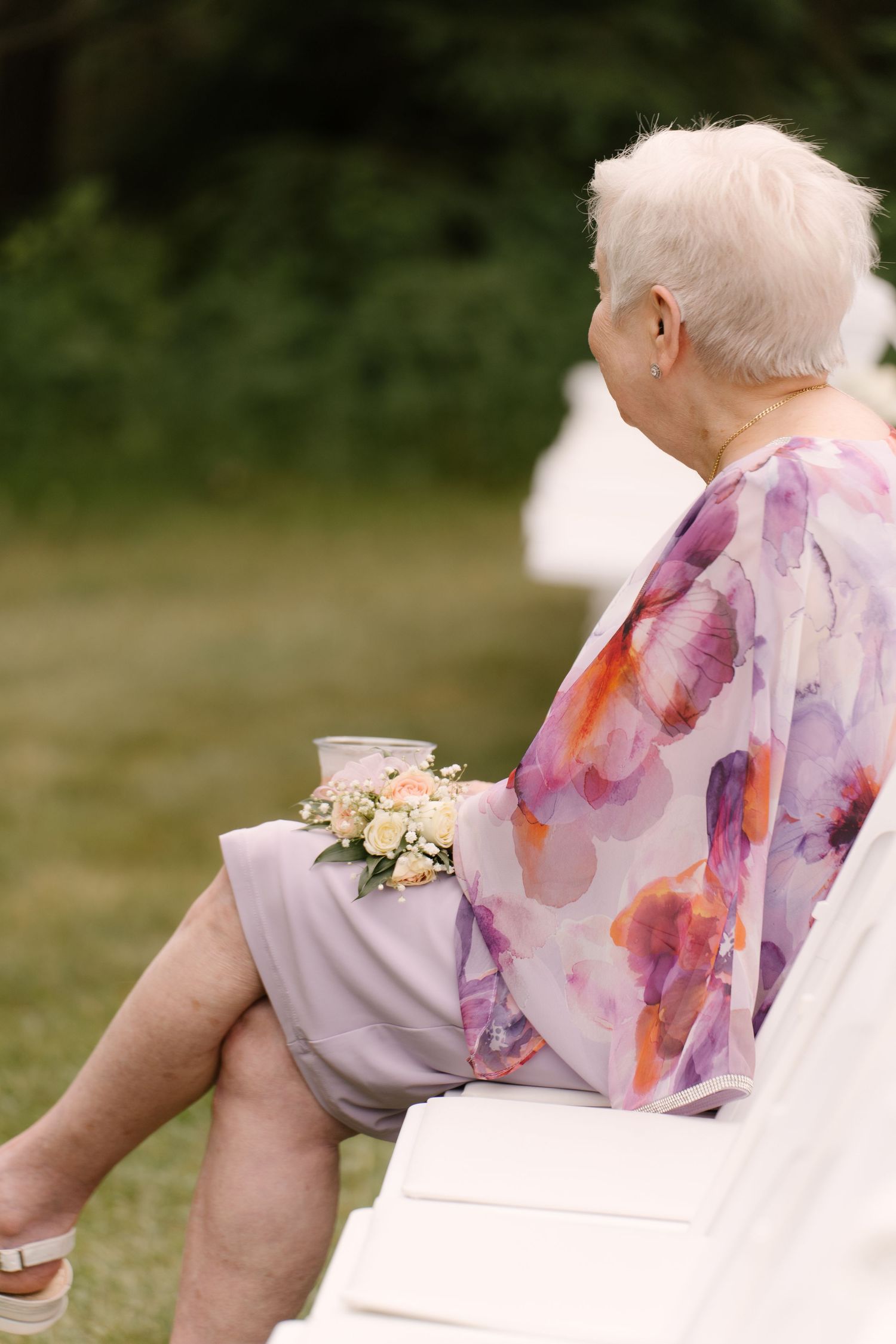 Elderly wedding guest in a floral dress with a wrist corsage sits peacefully at an outdoor ceremony in Guelph.