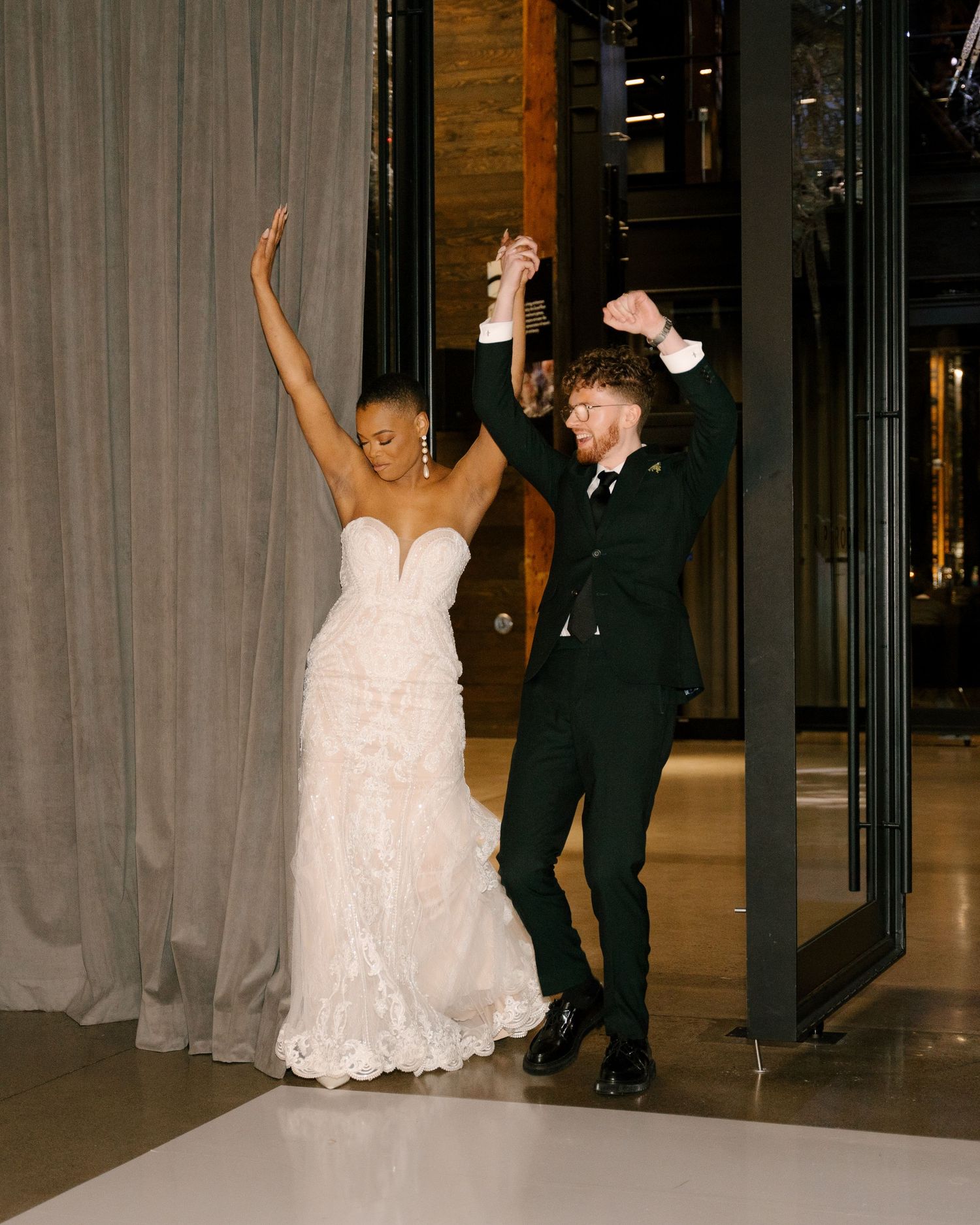 Joyful newlywed couple making grand entrance, bride in white lace gown and groom in dark green suit raising hands.