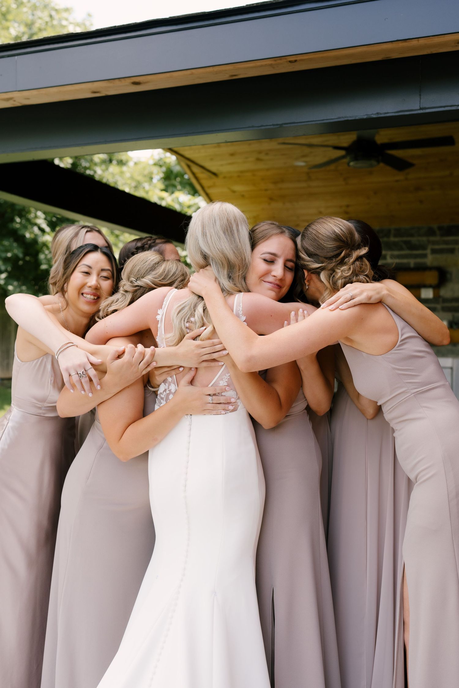 Bridesmaids in taupe gowns sharing a group hug with a bride in a white lace wedding dress outdoors.