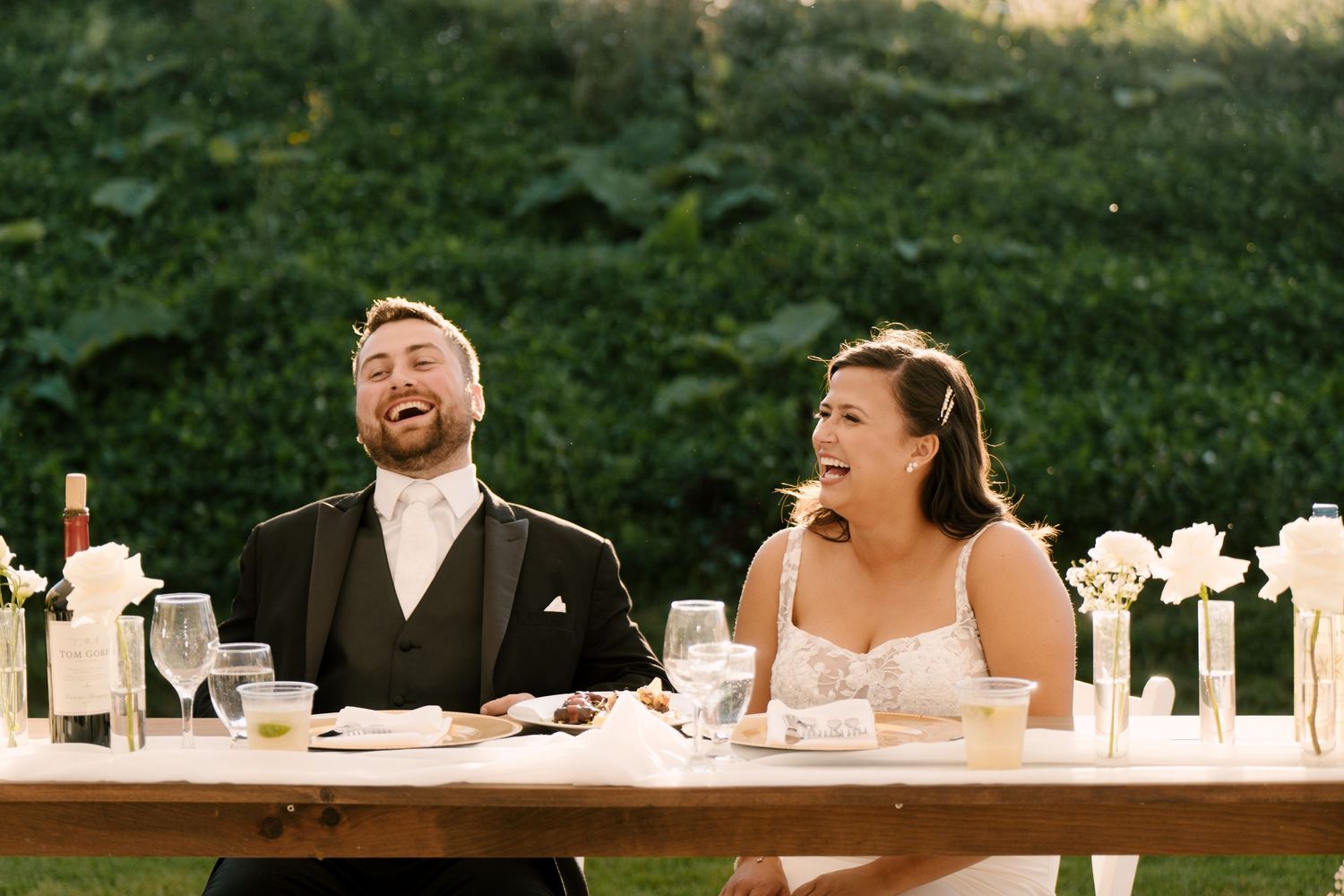 Bride and groom laughing joyfully at sweetheart table during outdoor wedding reception with white floral arrangements at Wakefield Estates.