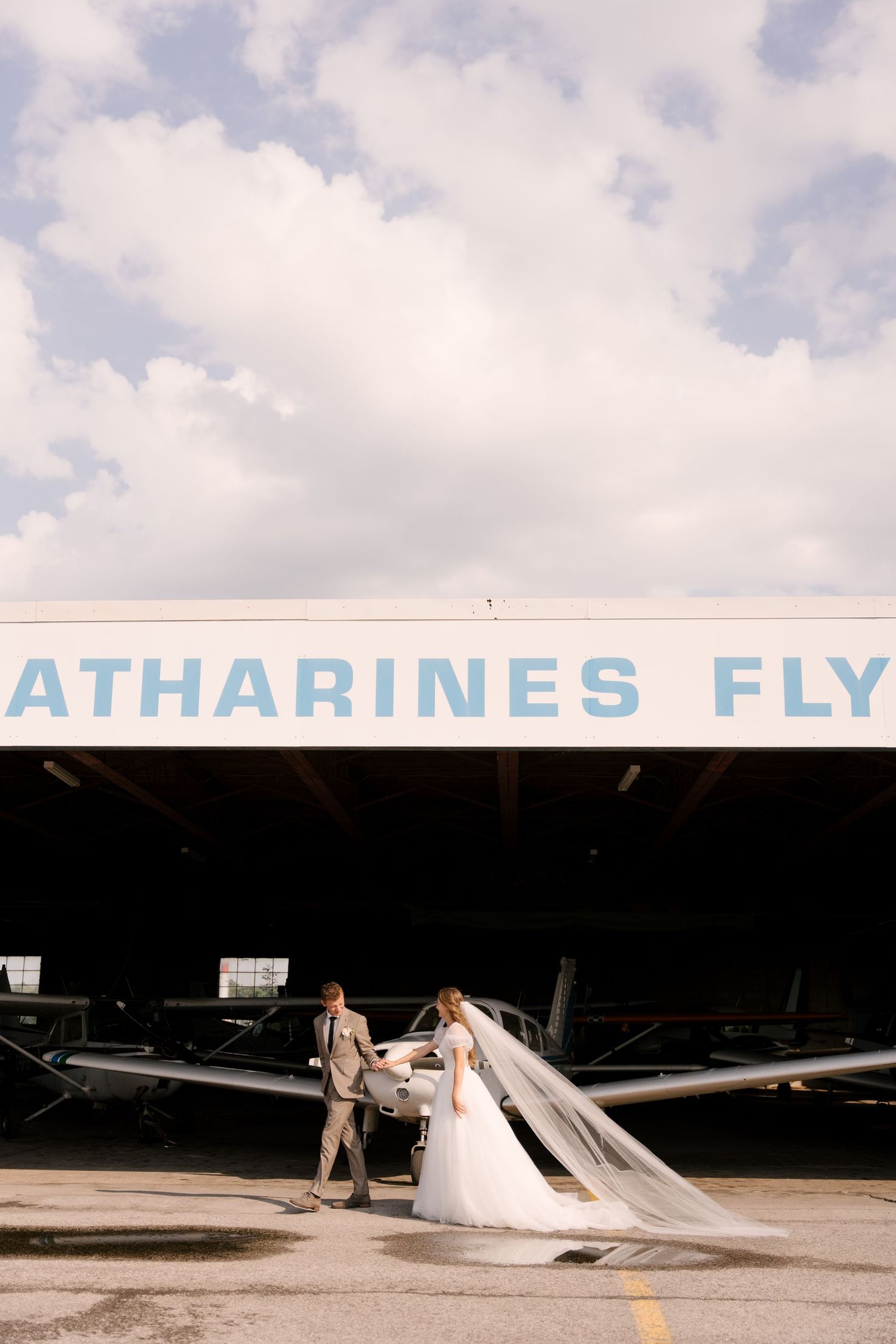 Bride with flowing veil and groom pose in front of St. Catharines Flying Club hangar with small planes.