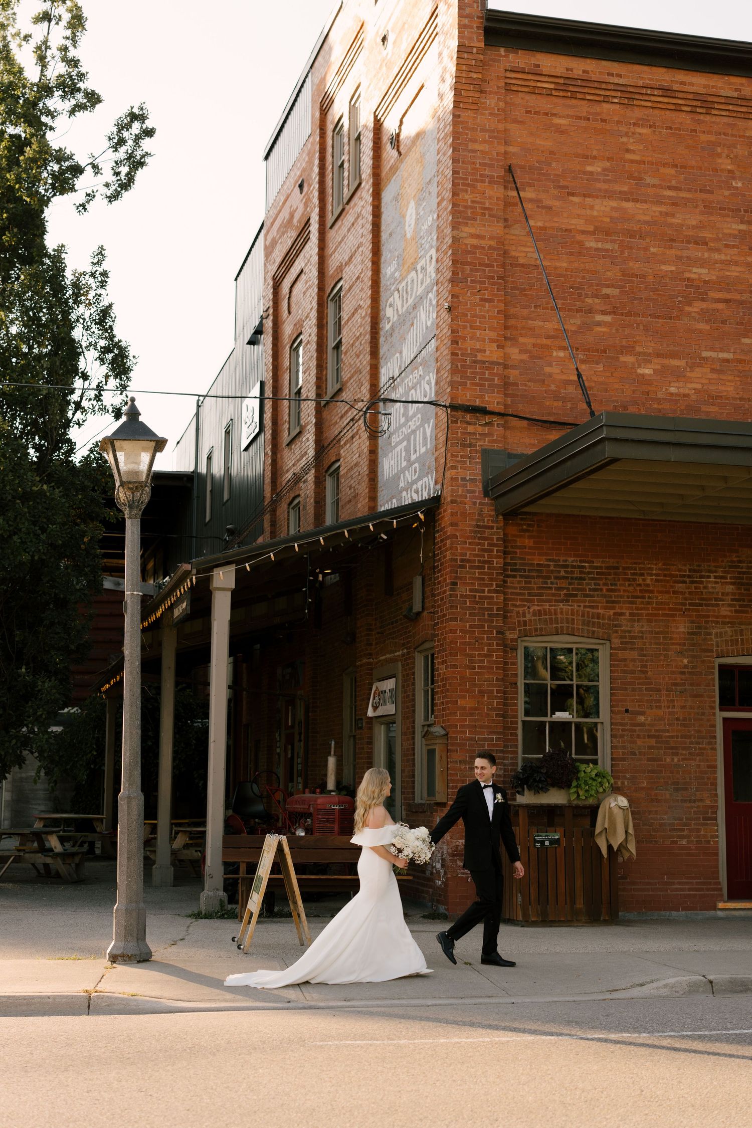 Bride and groom walk hand in hand past a rustic red brick building on their wedding day.