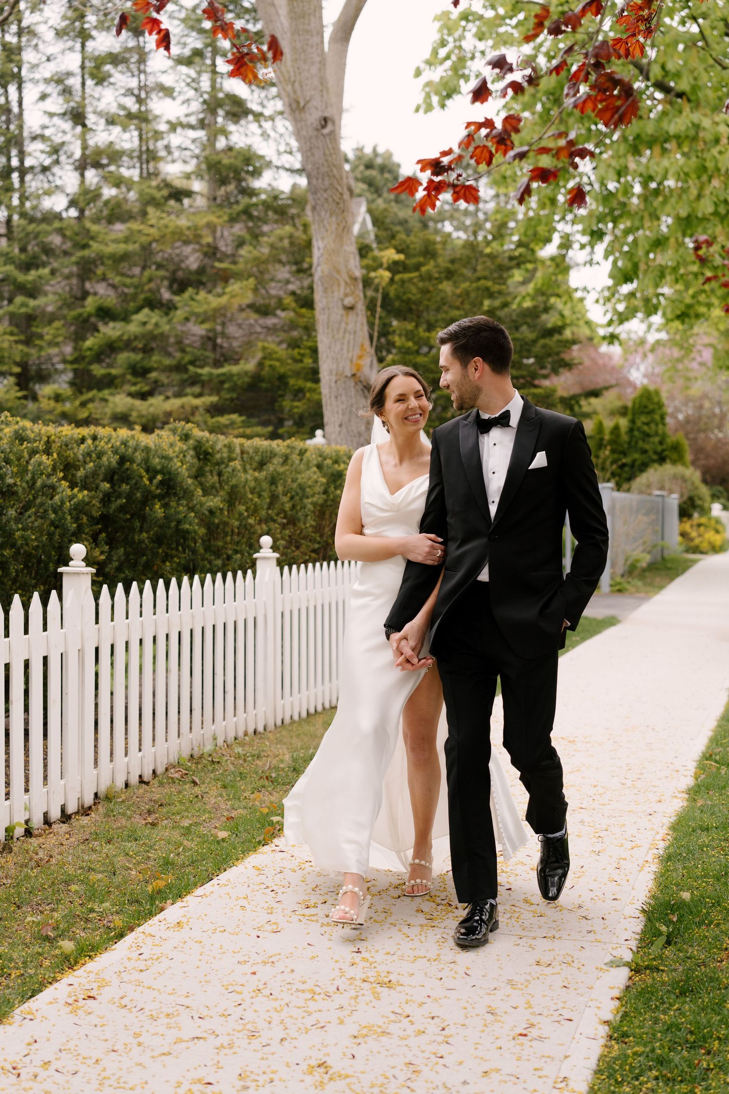 Bride and groom walk hand in hand along white picket fence path beneath autumn foliage.