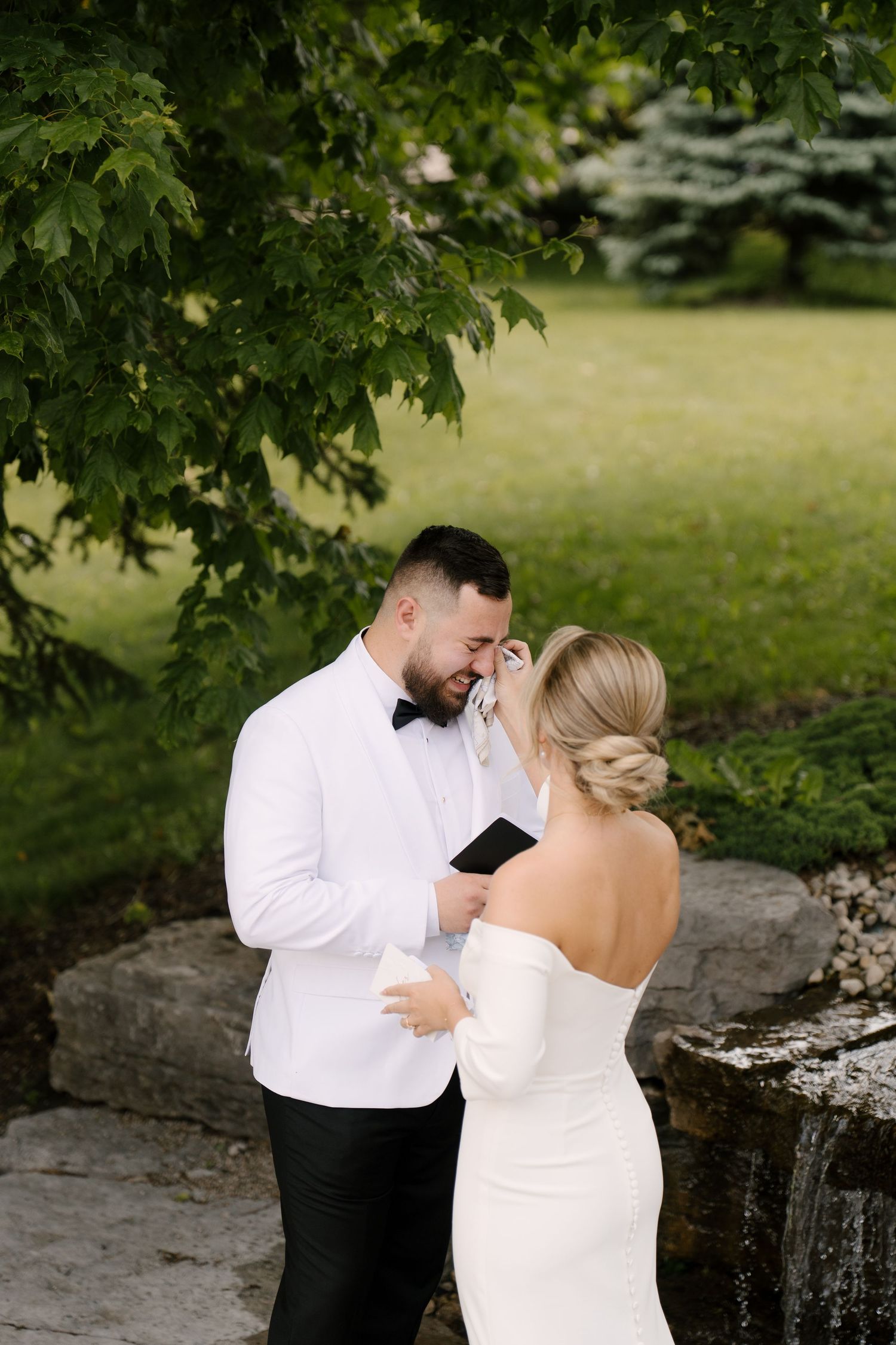 Emotional groom in white tuxedo wiping bride's tear during heartfelt first look in a lush green garden in Cambridge.