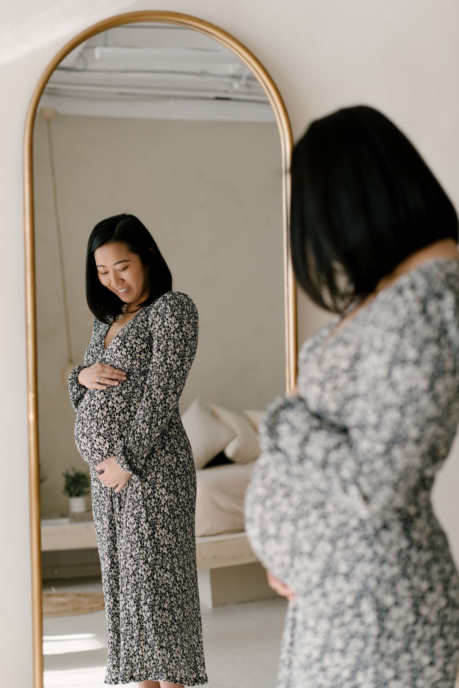 Pregnant Asian woman in floral dress admires her baby bump in a large gold-framed mirror.