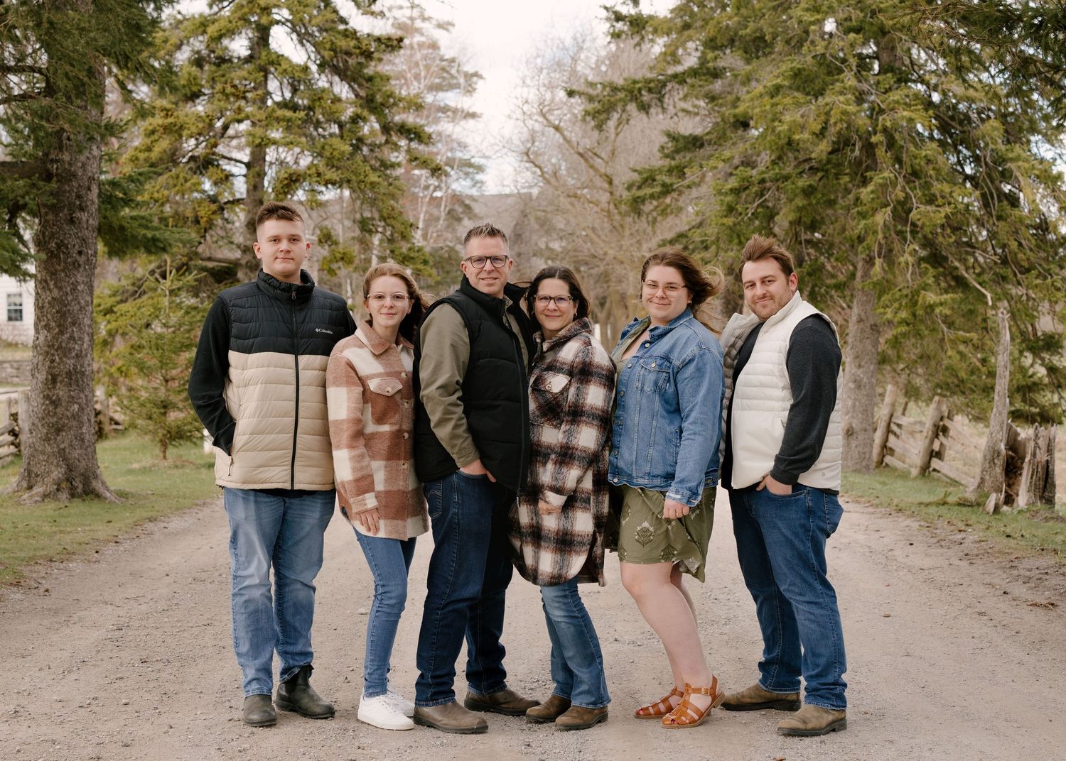 Family of six poses together smiling on a gravel path surrounded by tall evergreen trees outdoors.