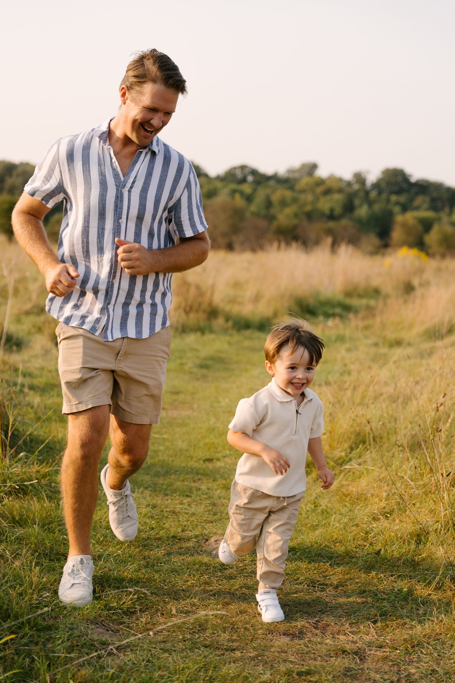 Laughing father chases his giggling young son along a sunlit grassy field path at golden hour.