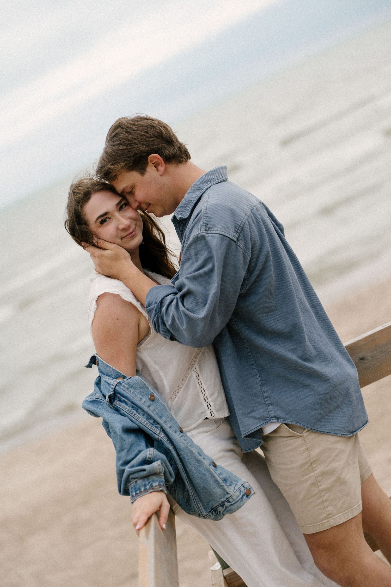 Romantic couple embracing on a pier by the ocean, woman smiling as man nuzzles her cheek tenderly.