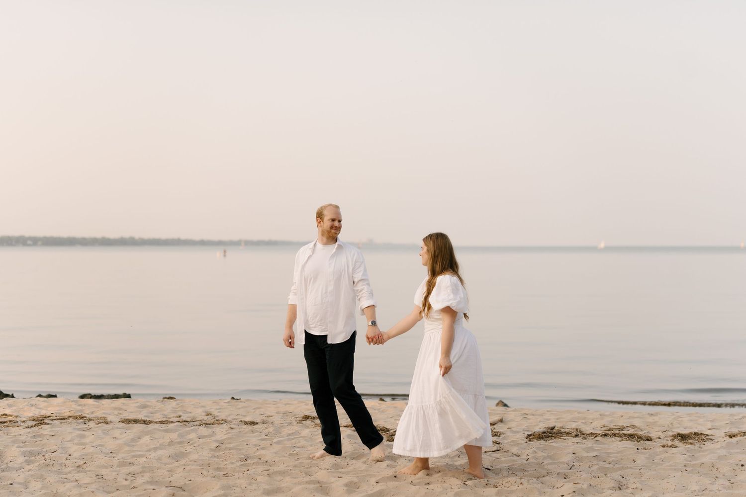 Couple holding hands and walking along the shoreline at sunset, dressed in white on a sandy beach.