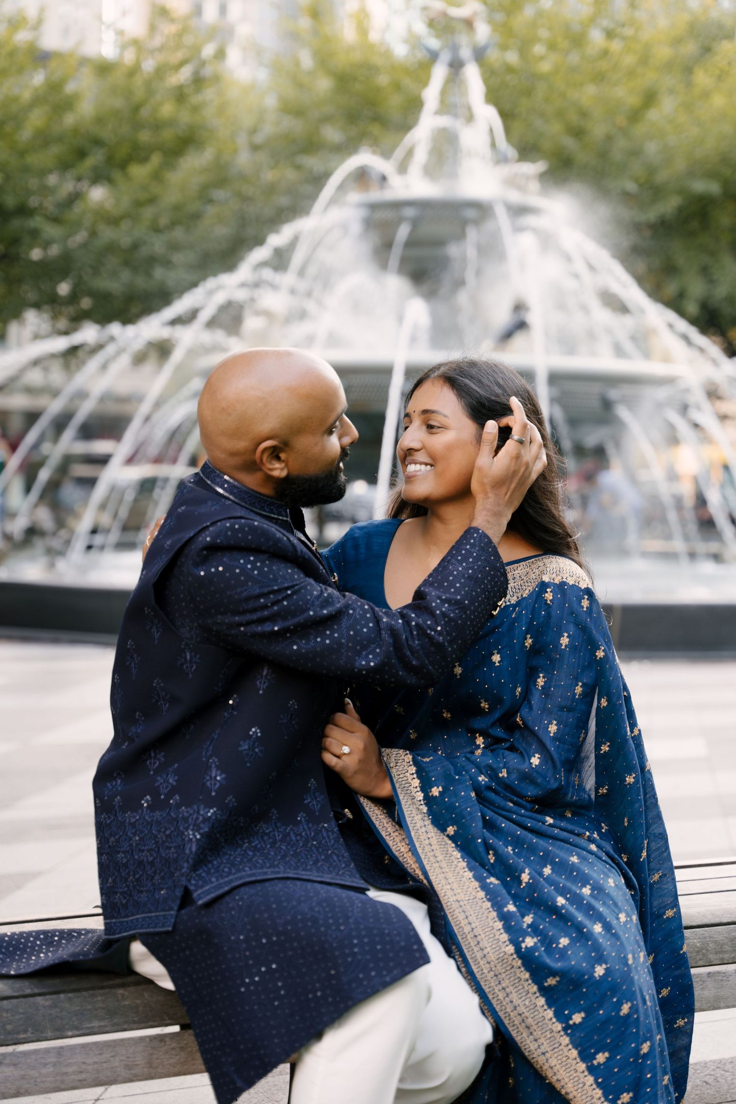 A couple in matching navy blue Indian attire share a romantic moment by a fountain.