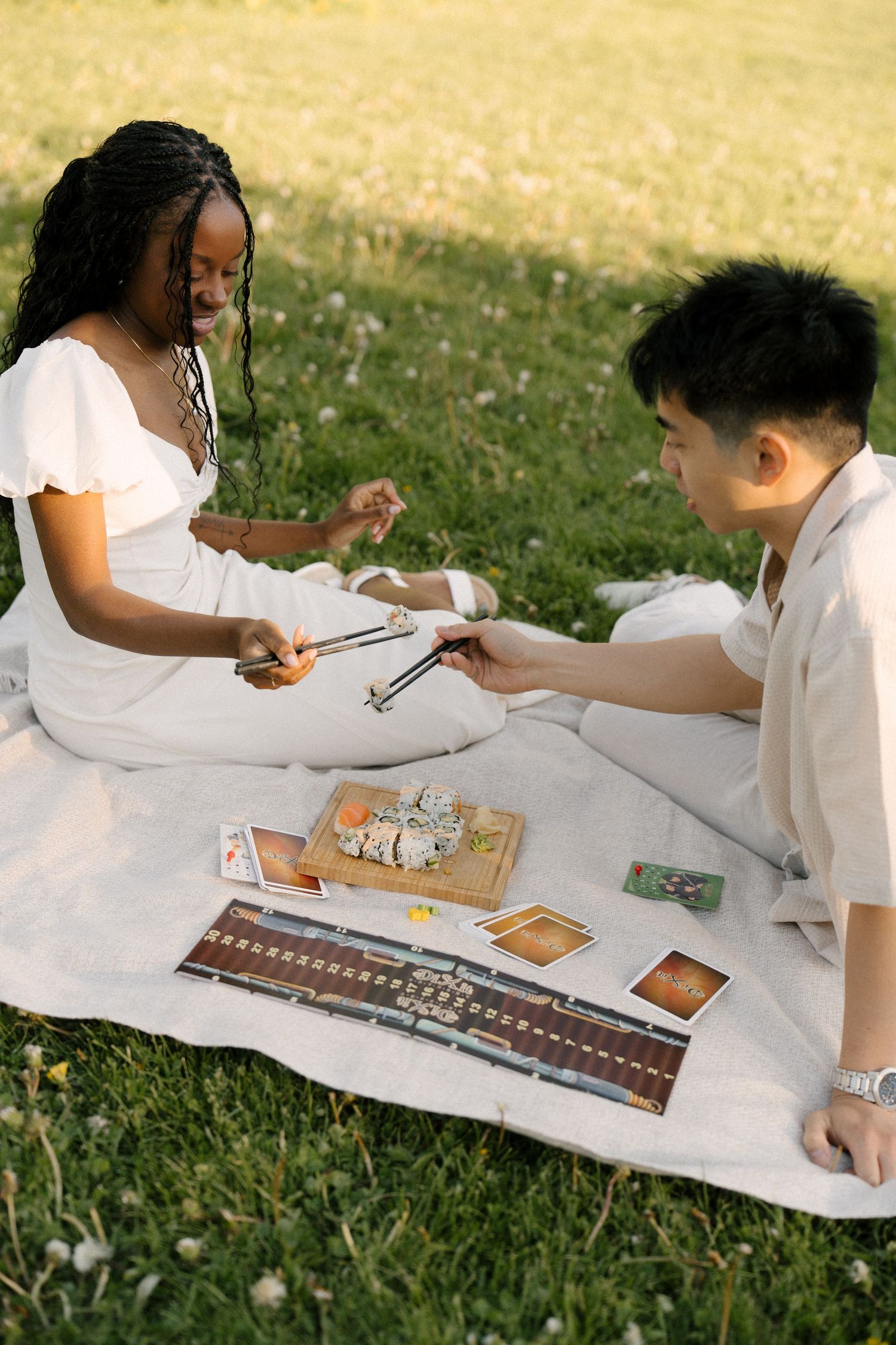 Couple sitting on blanket in sunny park sharing sushi with chopsticks during outdoor picnic.