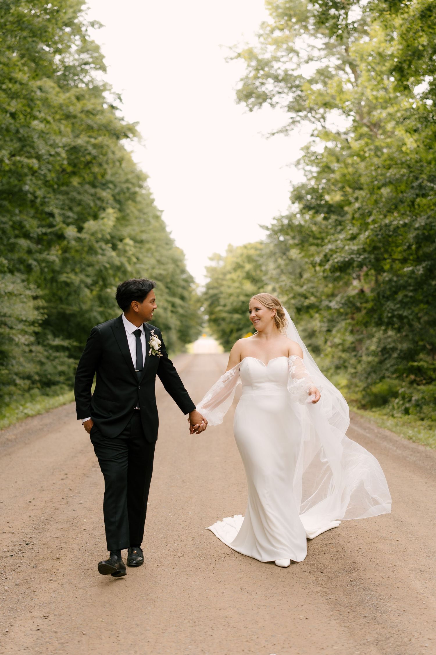 Bride and groom holding hands walking down a tree-lined dirt road, bride's veil flowing in the breeze.