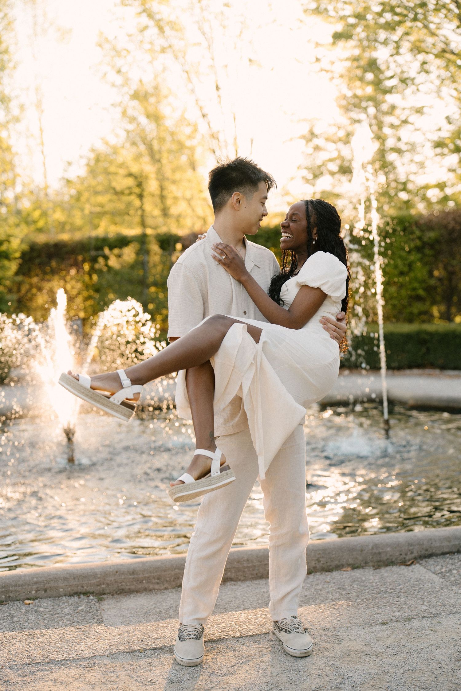 Man lifting woman in white dress near garden fountain at golden hour.