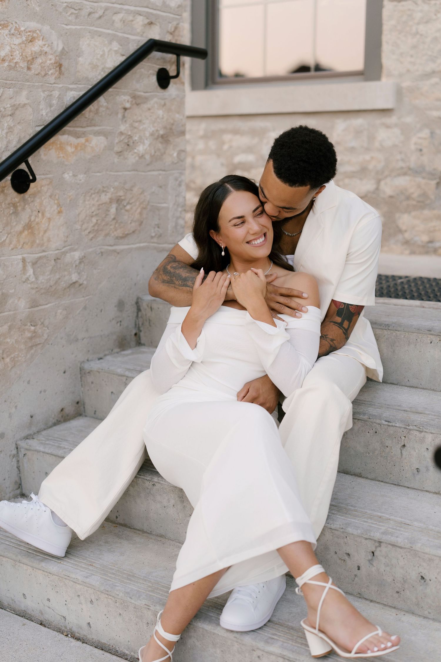 Couple in white outfits sitting on stone steps, sharing a romantic embrace and kiss.