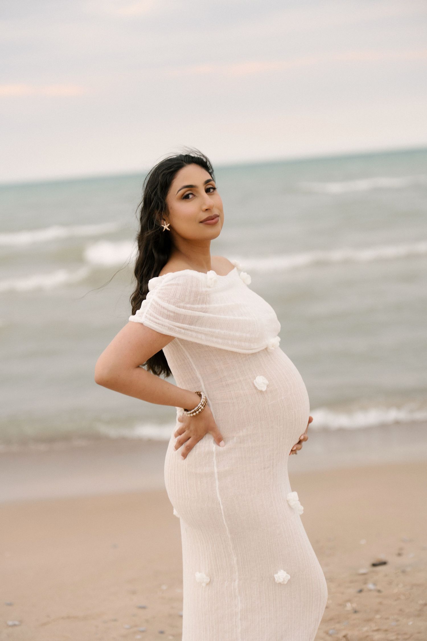 Elegant pregnant woman in off-shoulder white dress cradling baby bump on a breezy beach at dusk.