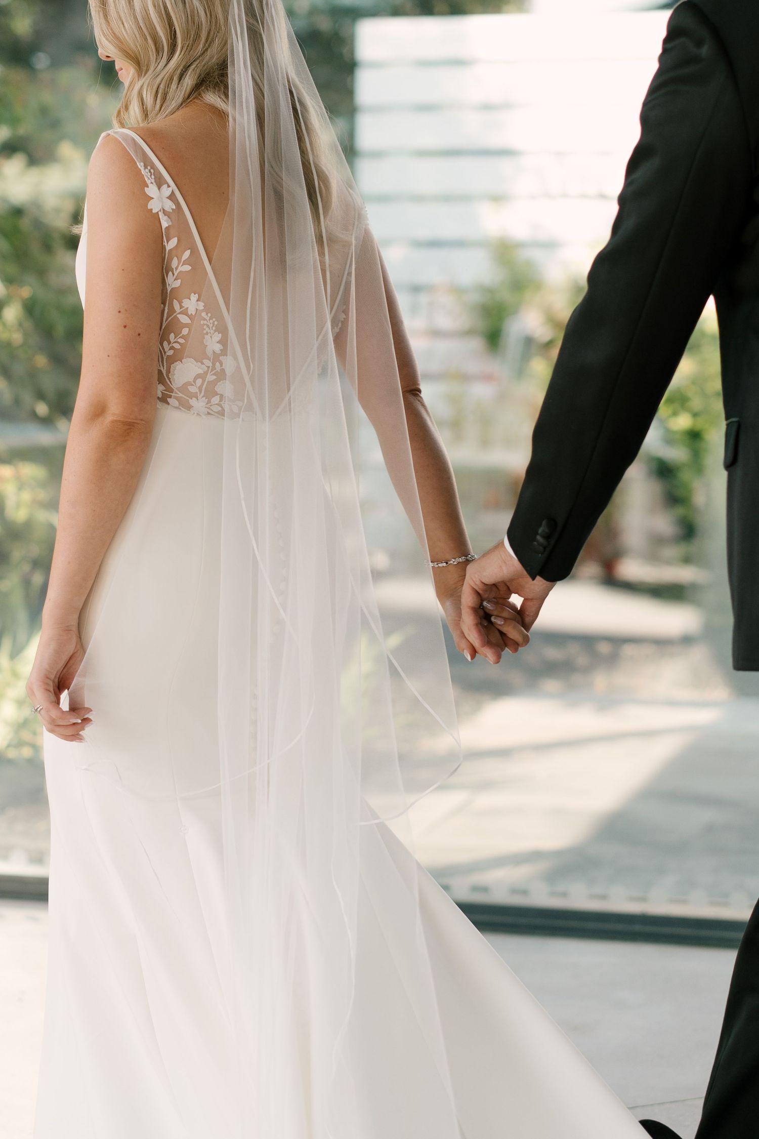 Rear view of a bride in a lace-back wedding gown and veil holding hands with her groom indoors.