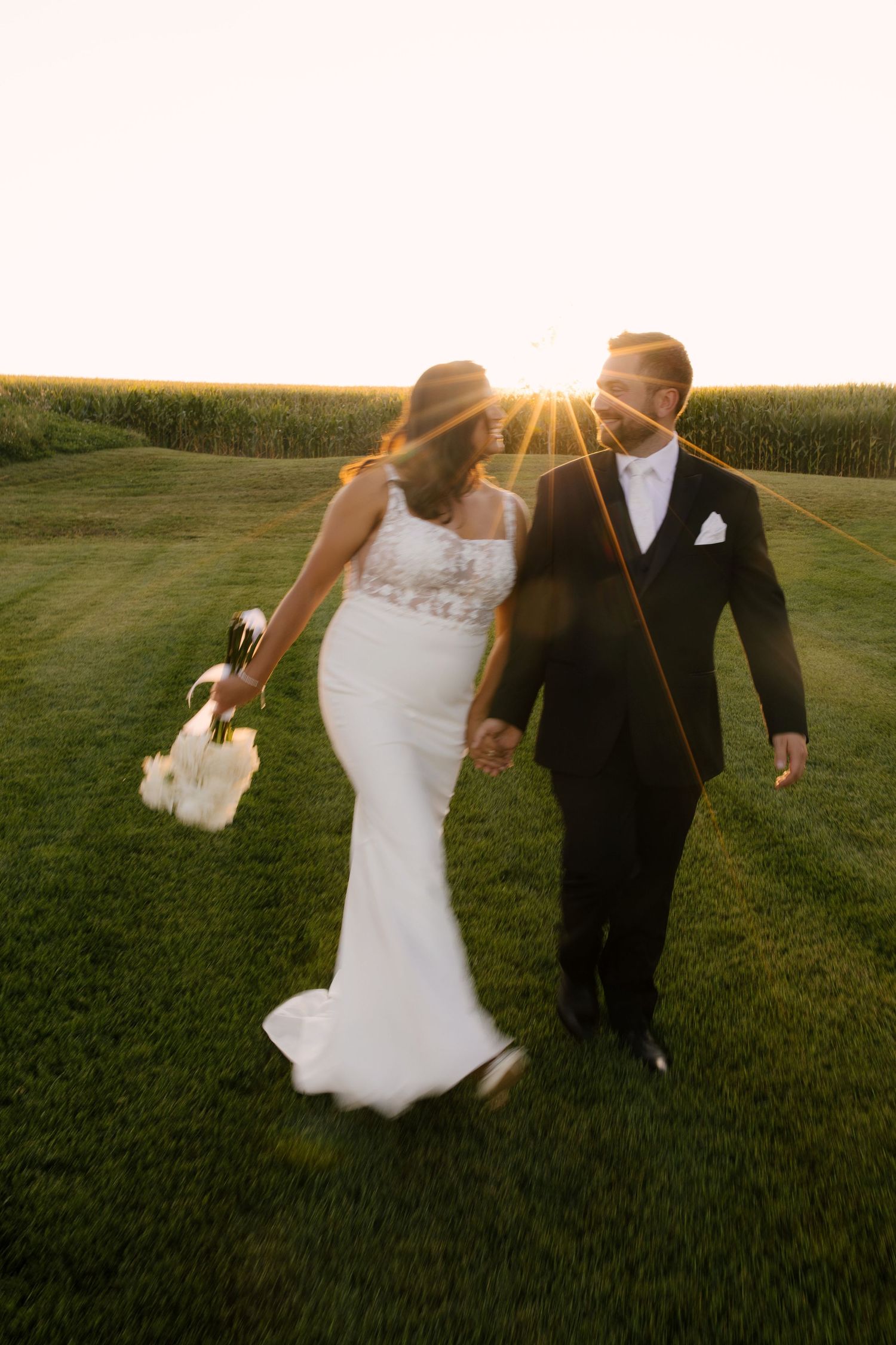 Newlyweds walking hand in hand through green fields with stunning golden sunset behind them at Wakefield Estates.