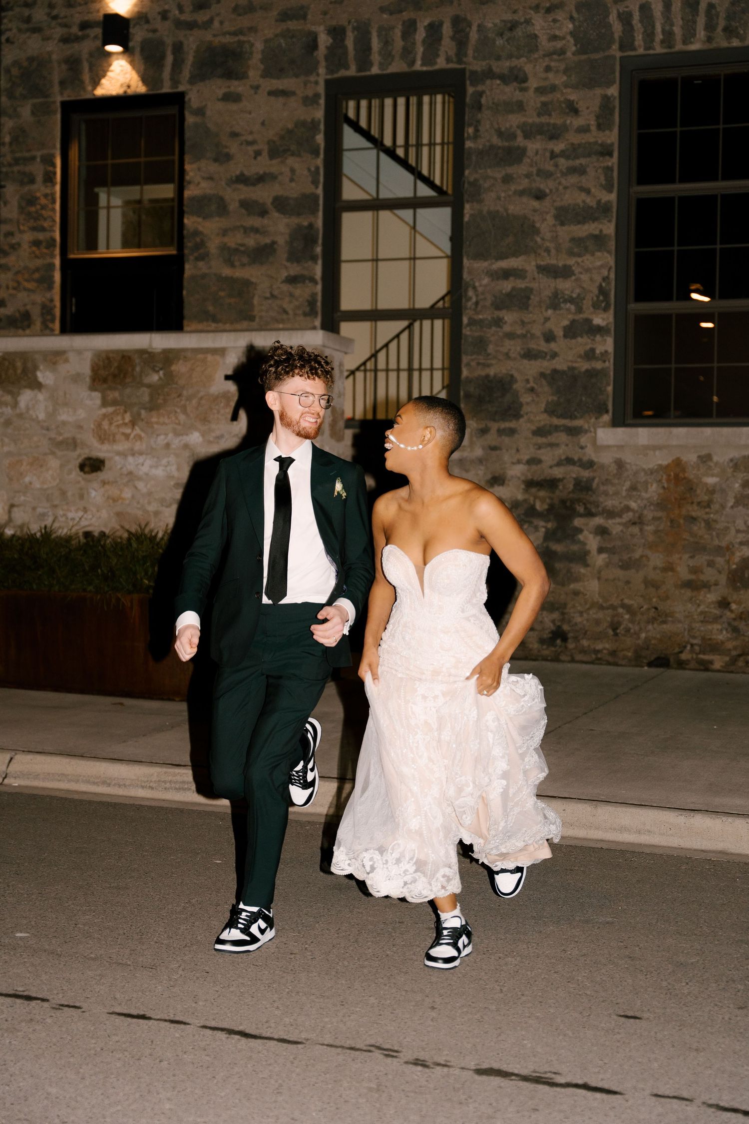 Bride in lace gown and groom in dark suit running joyfully together outside at night wearing sneakers.