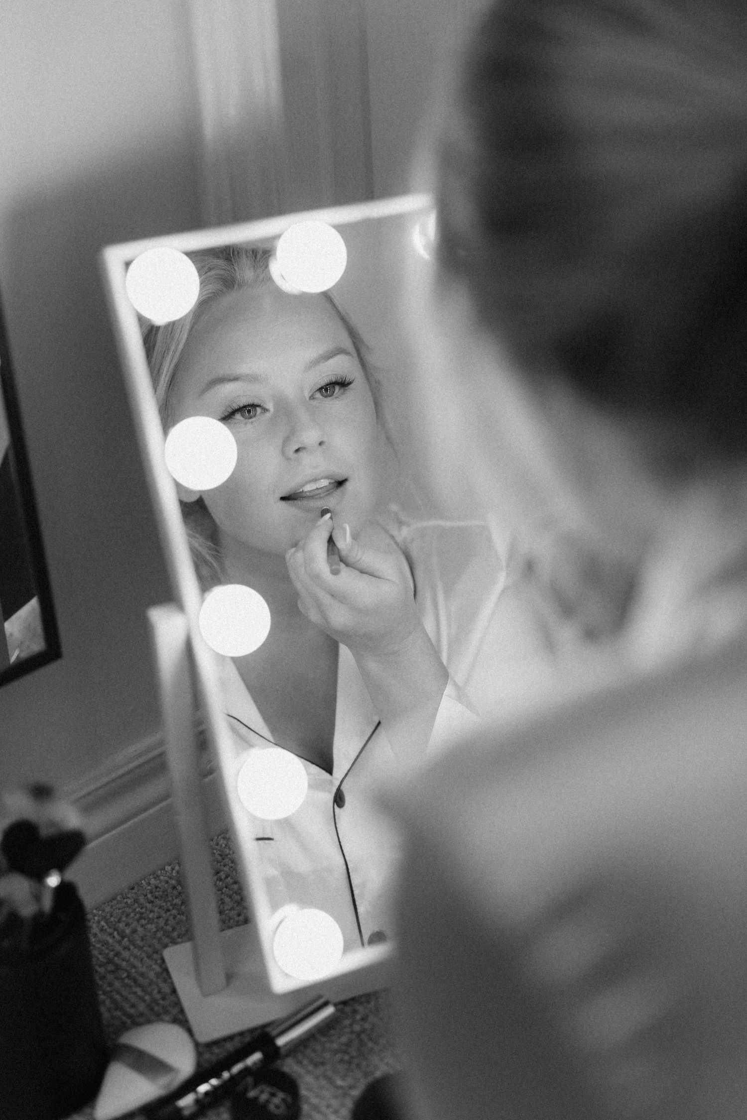 Bride applying lipstick at a Hollywood vanity mirror with glowing bulbs in black and white.