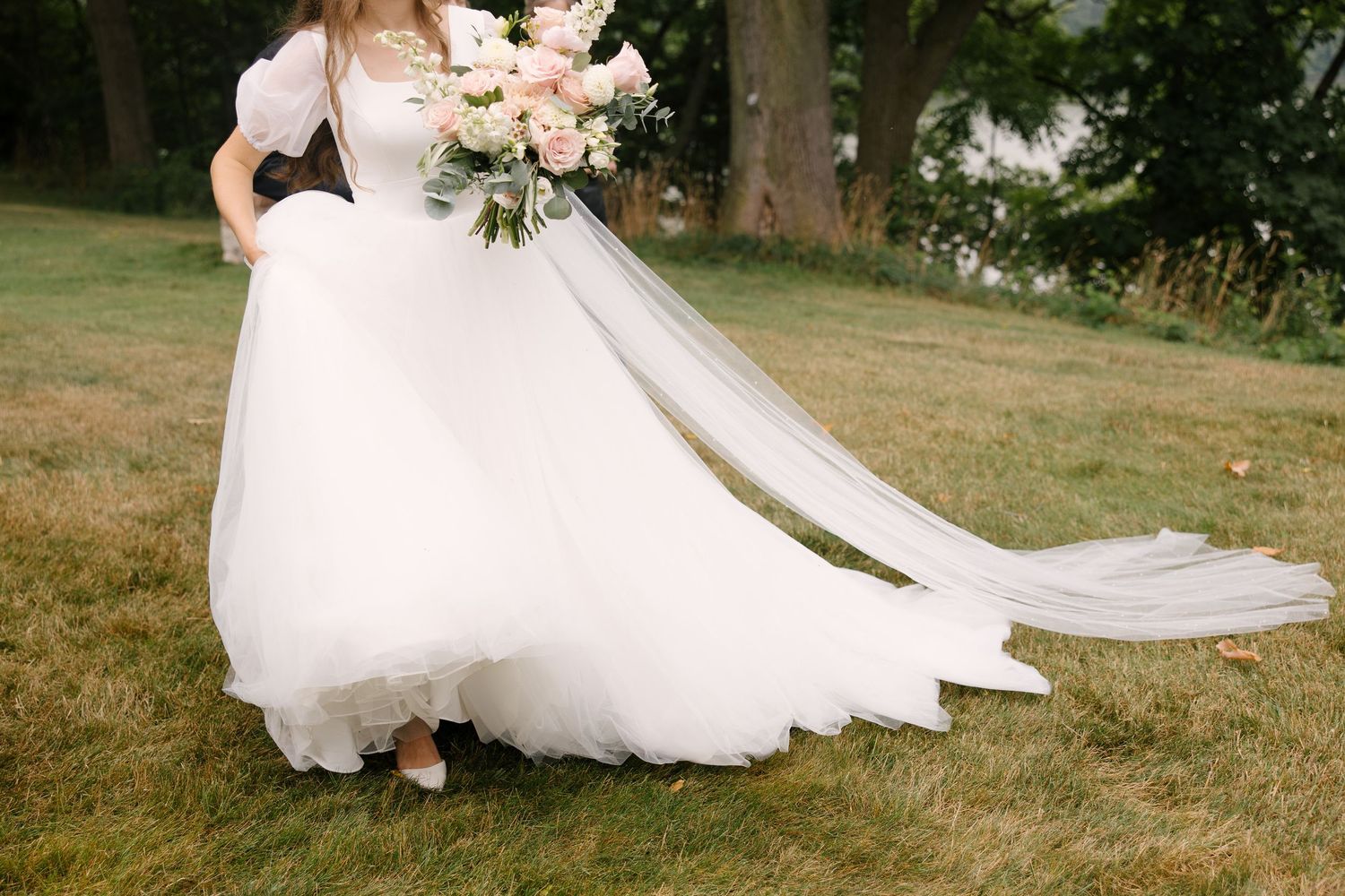 Bride in a white ball gown with flowing veil walks across a green lawn holding a blush bouquet.