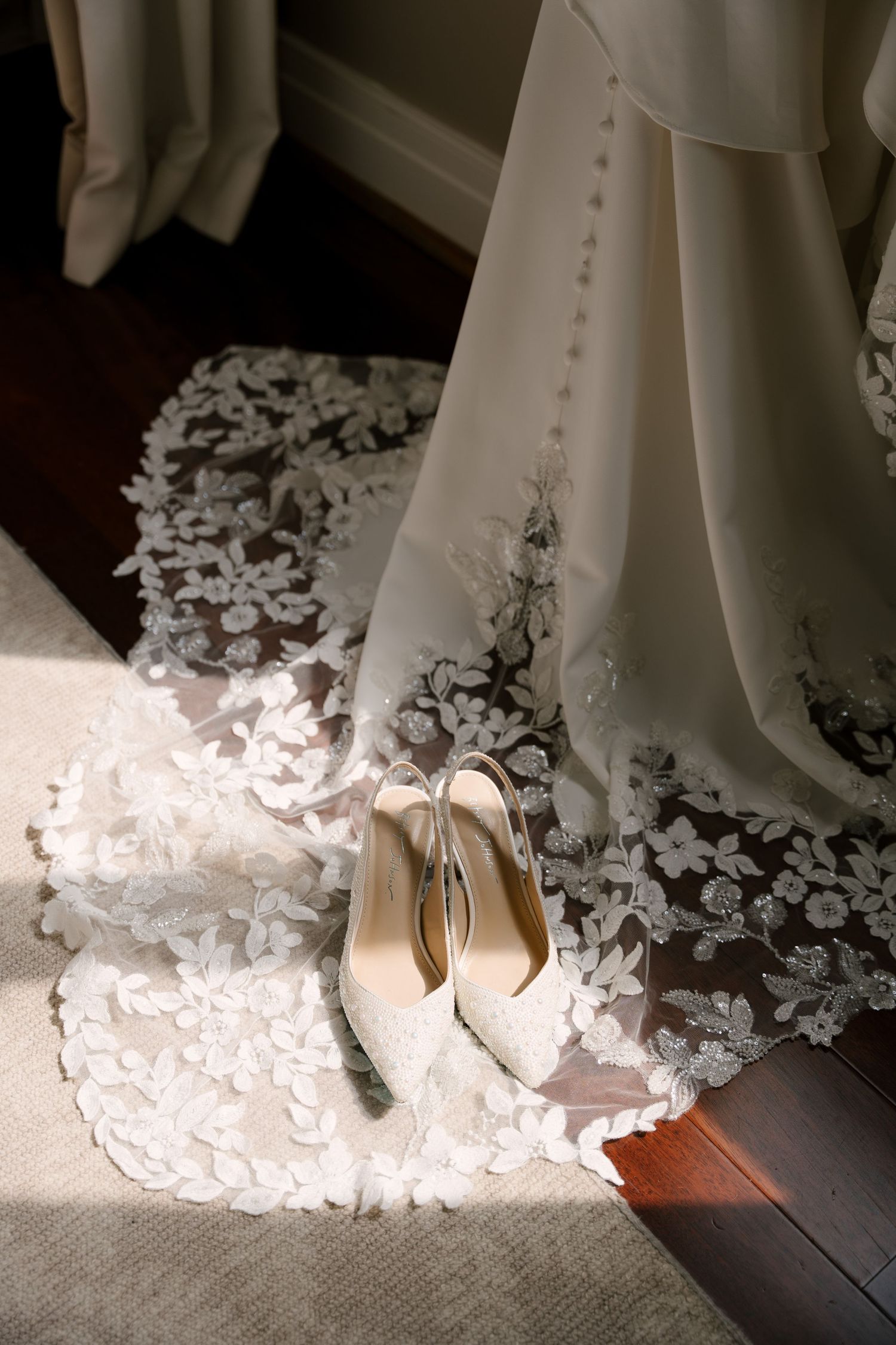 Elegant bridal wedding dress with floral lace train displayed with ivory satin heels on hardwood floor in natural light.