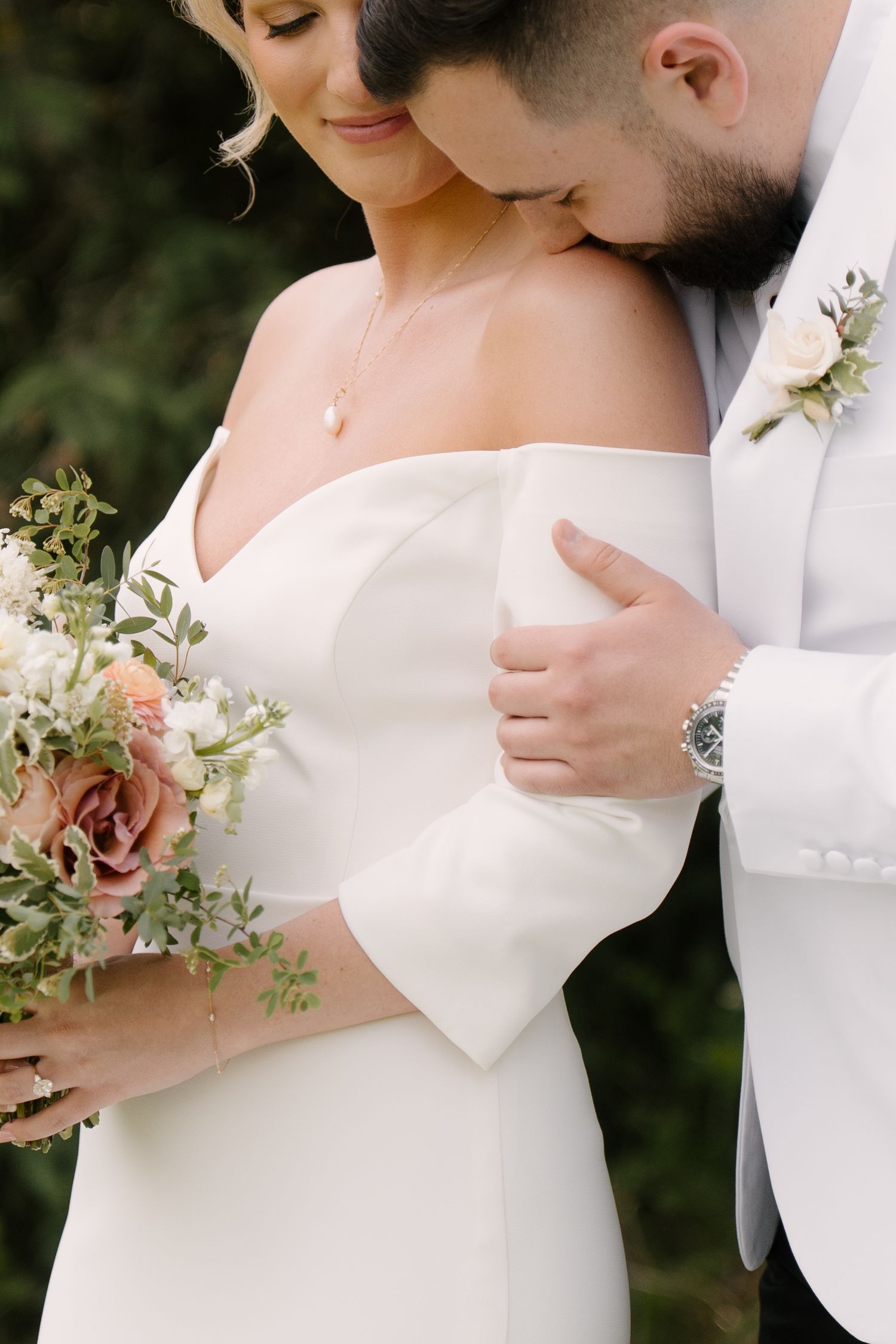 Groom in white tuxedo kissing bride's shoulder as she holds a blush and white floral wedding bouquet.