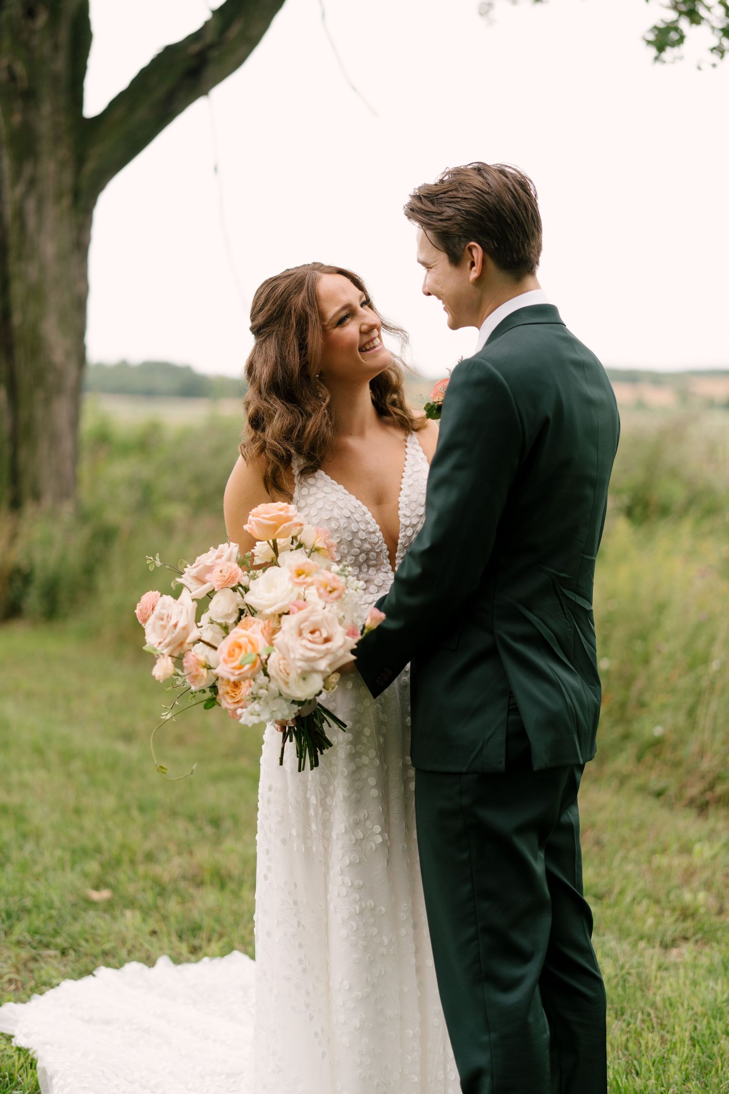 Happy bride and groom sharing romantic moment under tree holding blush rose bouquet outdoors.
