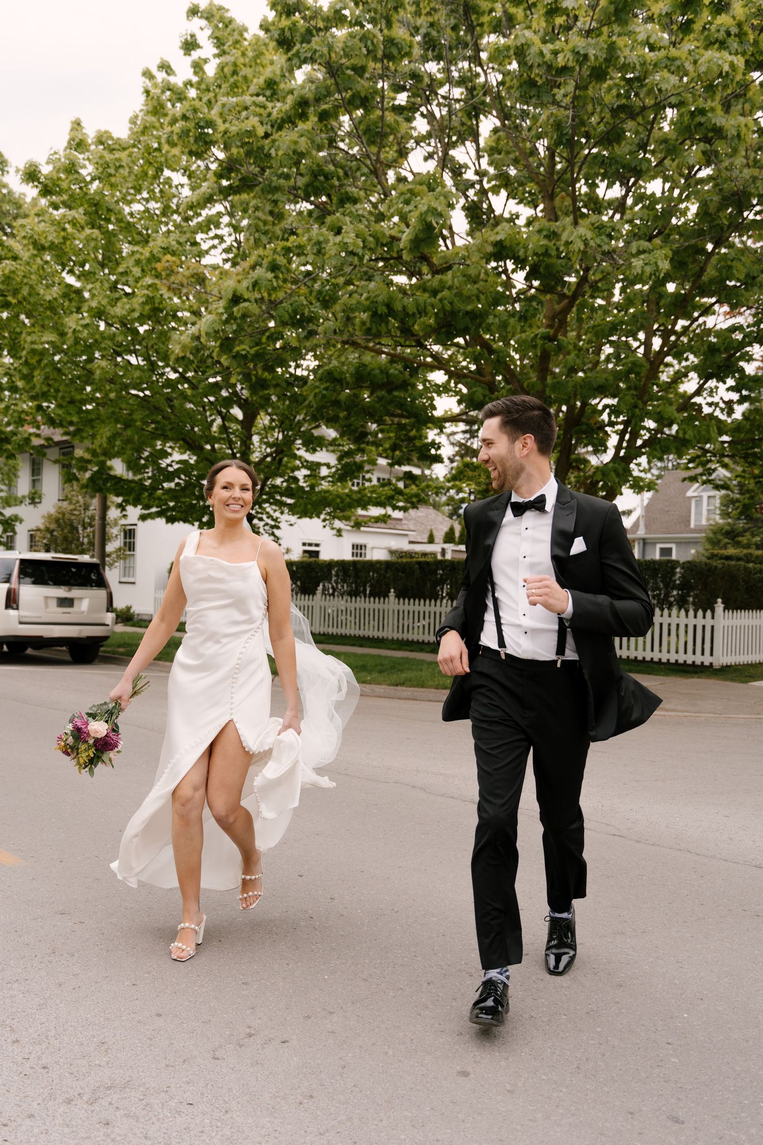 Joyful bride and groom walk across street, bride holding colourful bouquet in flowing white gown downtown Niagara-on-the-lake.