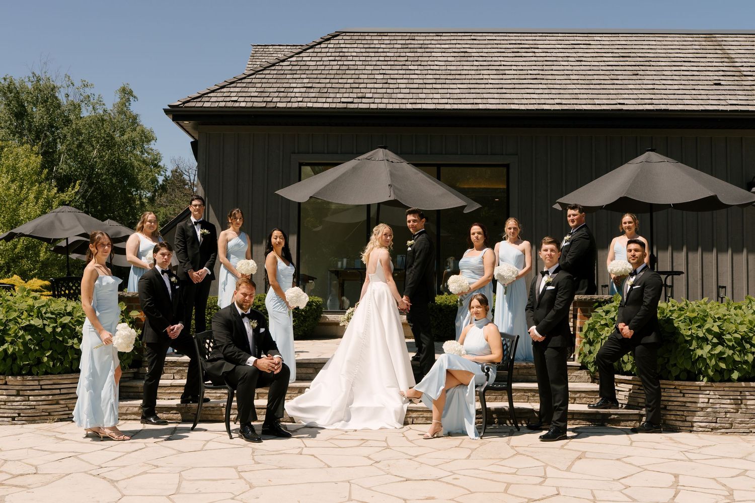 Full wedding party poses on stone patio outside elegant Roseville Estates wedding venue on sunny day.