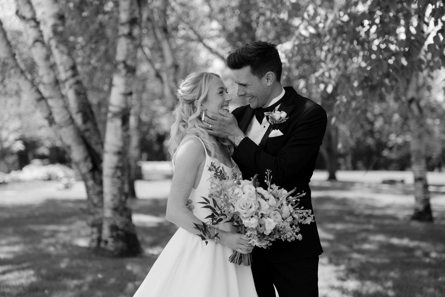 Newlywed couple shares a tender moment among birch trees, bride holding bouquet at Roseville Estates wedding venue.