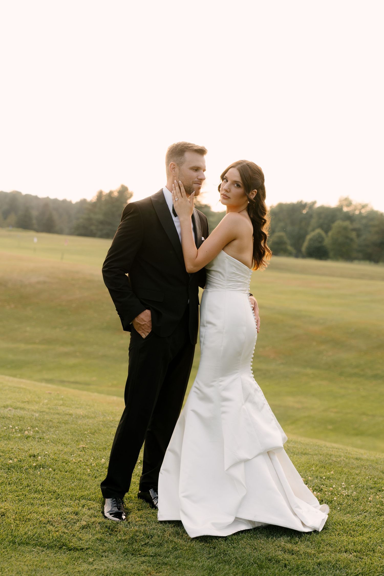 Bride in a strapless mermaid gown and groom in black tuxedo posing at sunset on a golf course.