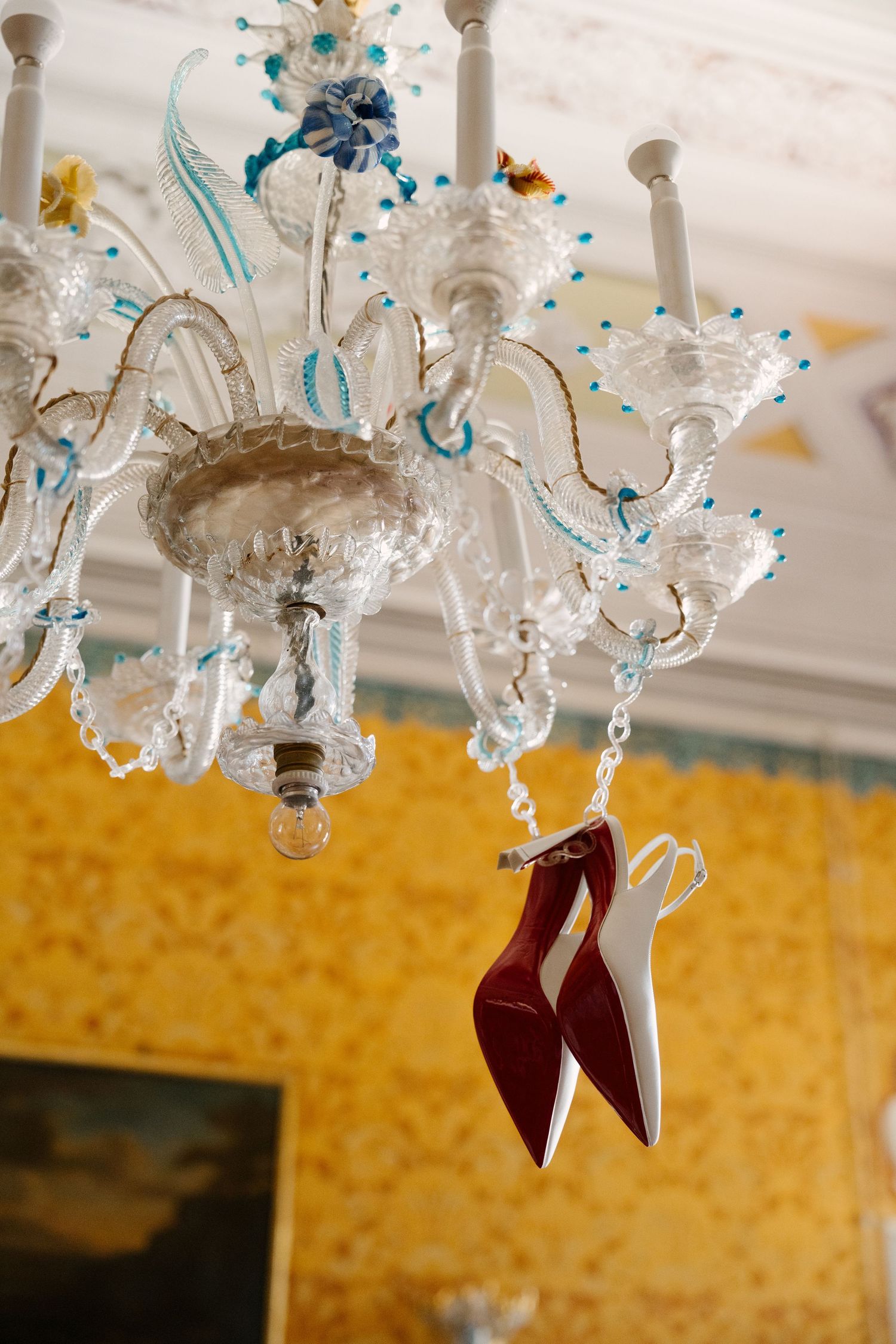 Red-soled bridal heels hanging elegantly from an ornate Murano glass chandelier against yellow wallpaper backdrop.