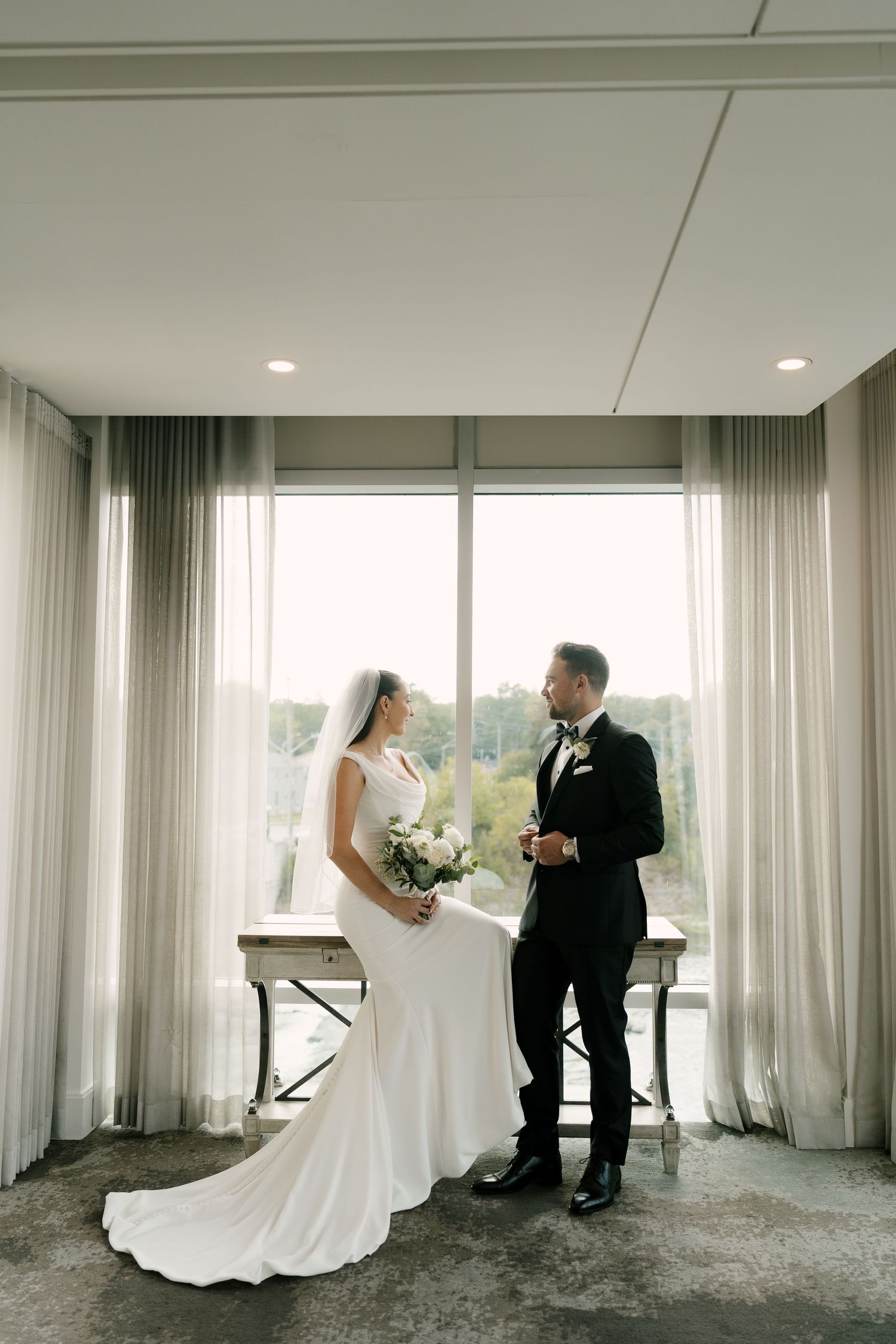 Elegant bride in white gown holds bouquet while groom in black tuxedo stands by bright curtained windows at Cambridge Mill.