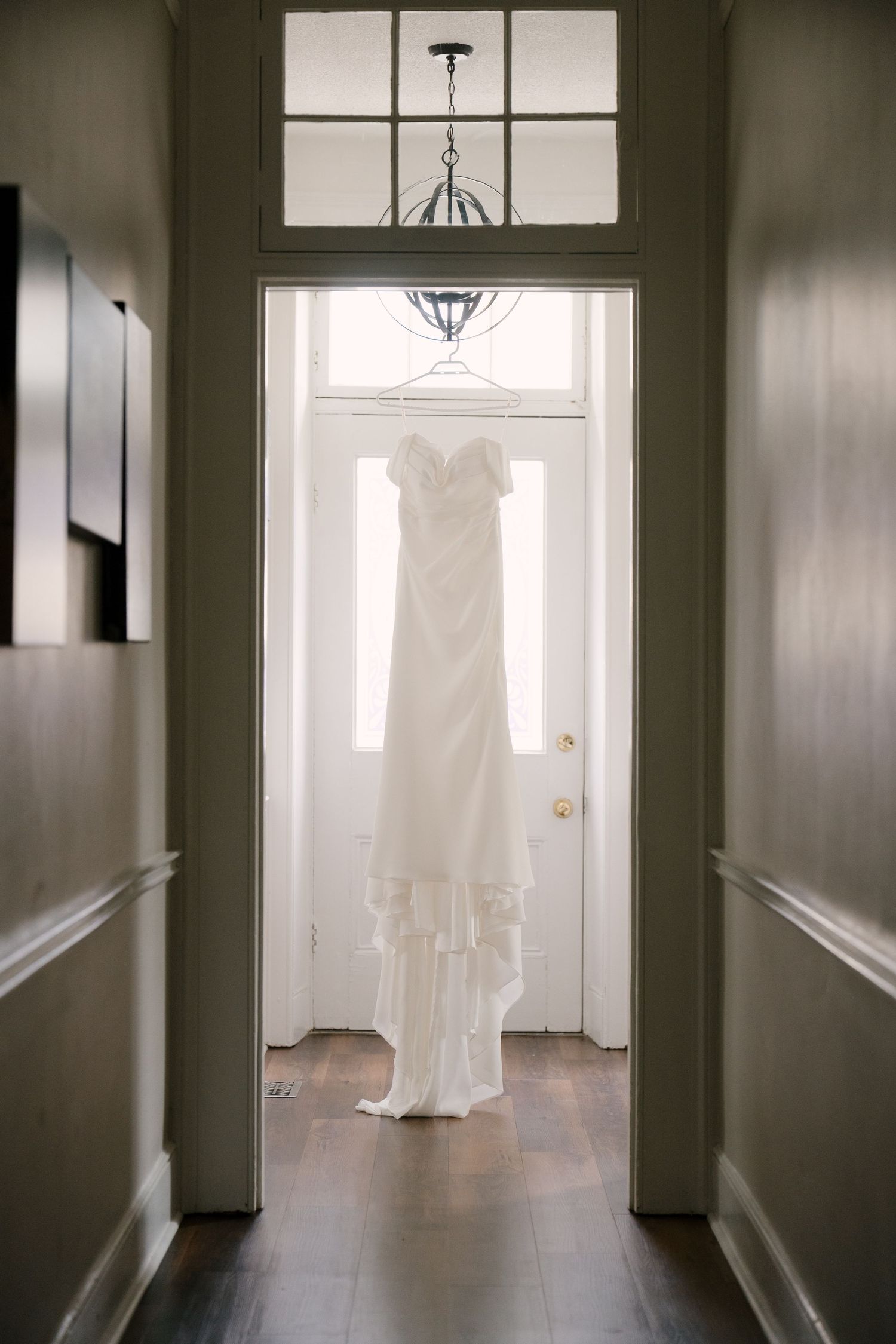 Elegant white wedding gown with ruffled hem hanging in sunlit doorway of classic home.